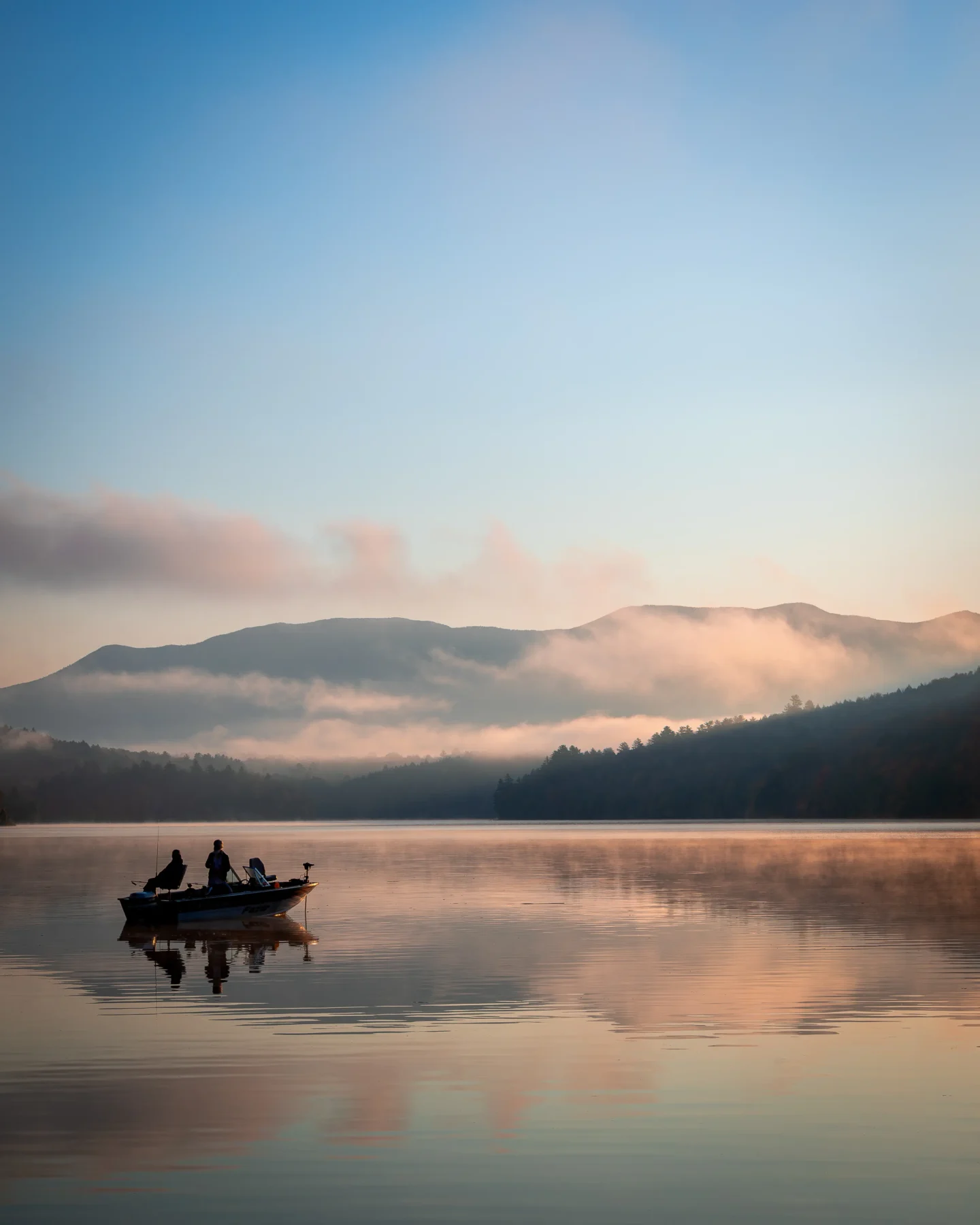silhouette of two people in a small boat in middle of a lake at sunrise time with fog coming off the lake and silhouette of a mountain range in the distance and soft blue sky above.