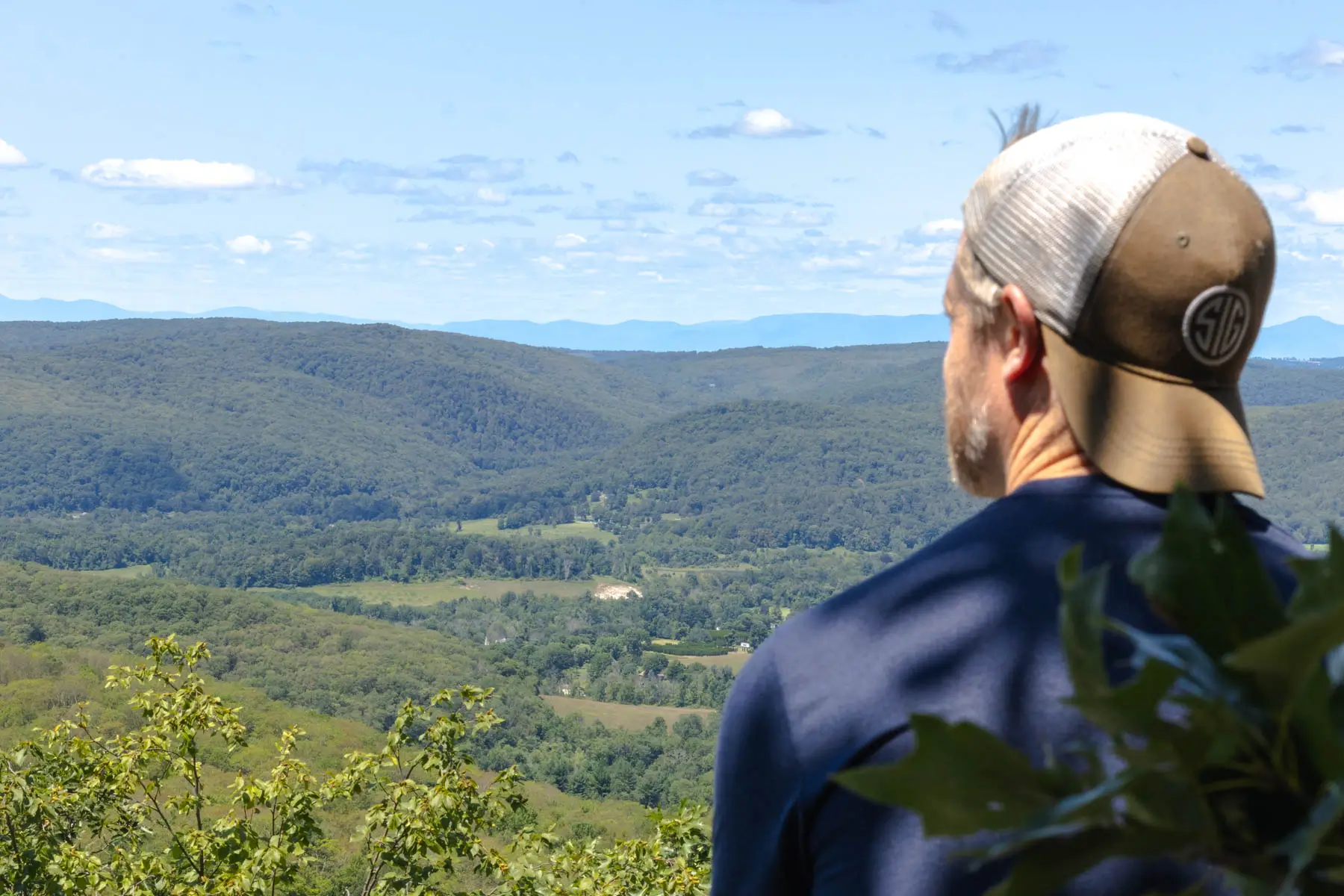 back of man sitting on top of hike with background baseball hat and rolling green hills and mountains in the distance behind him and soft blue sky with fluffy white clouds above.