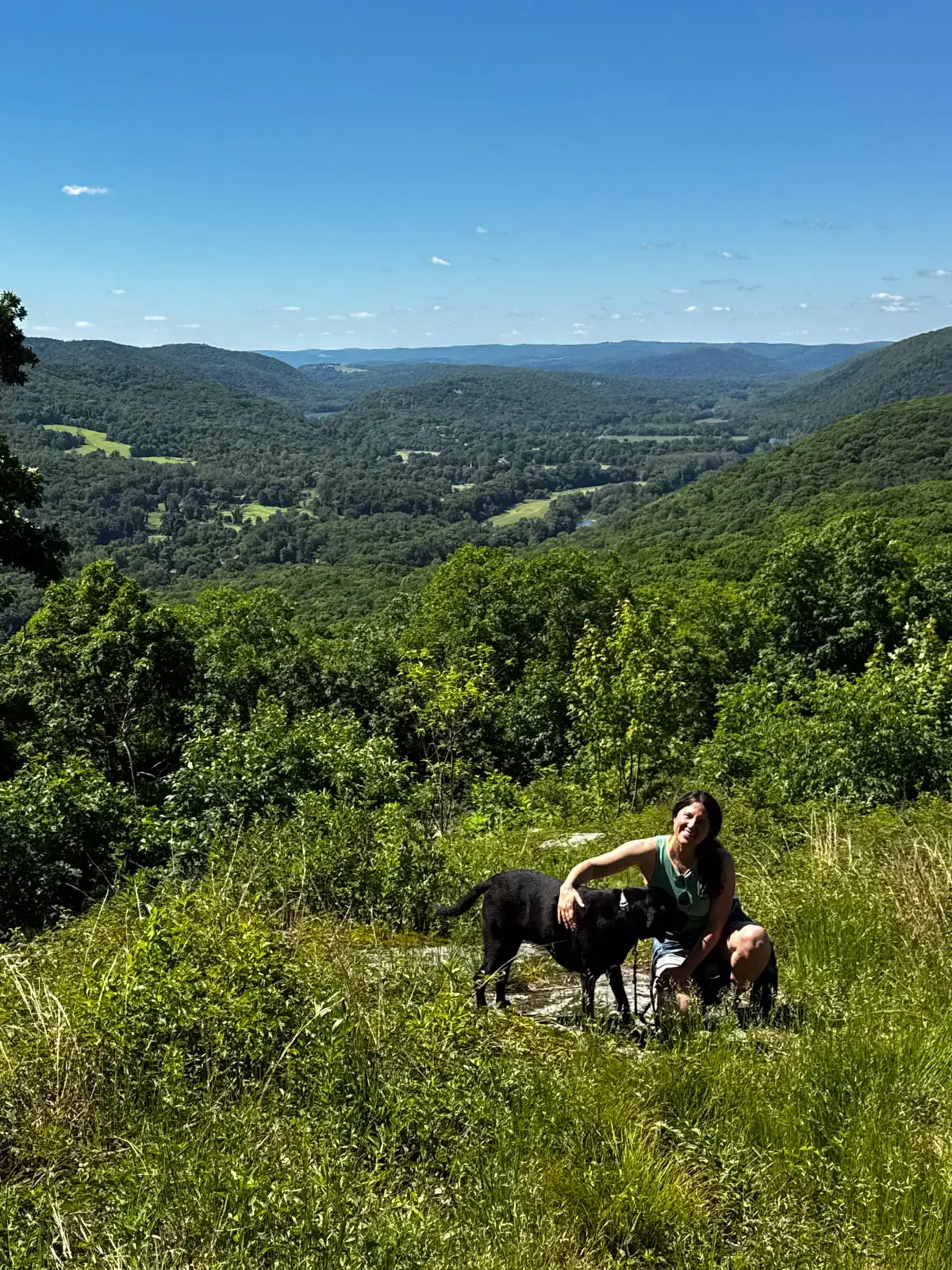 woman crouching down with black dog at top of hike in connecticut with rolling green hills in background and blue sky above.