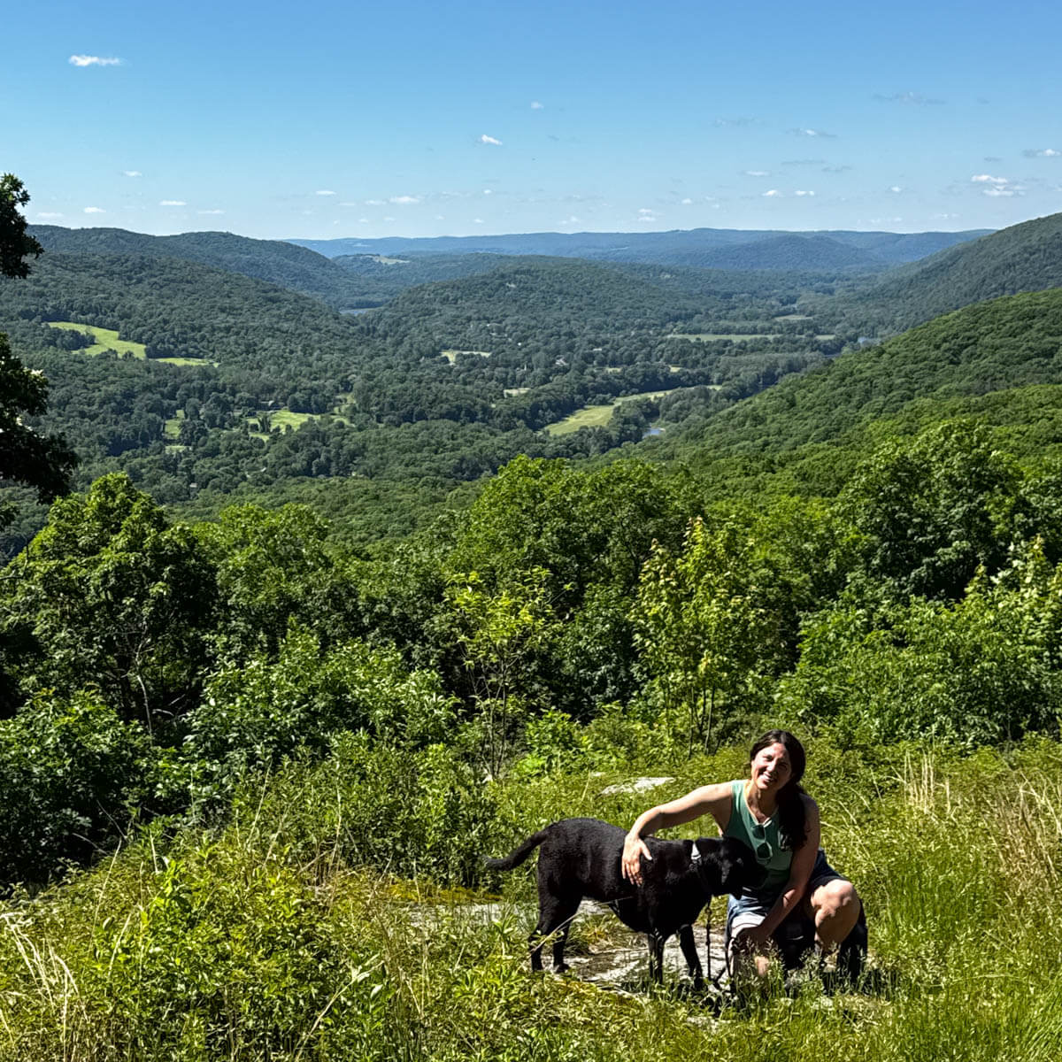 woman squatting down next to black dog on hike in connecticut in summer with rolling green hills in the distance.
