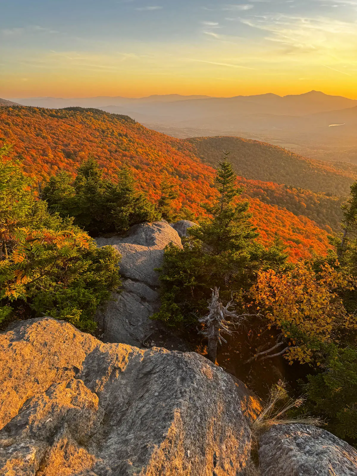 view from top of stowe pinnacle hike in fall at sunset time with view of bright orange colored mountains rolling in the distance and soft orange glow in the sky.