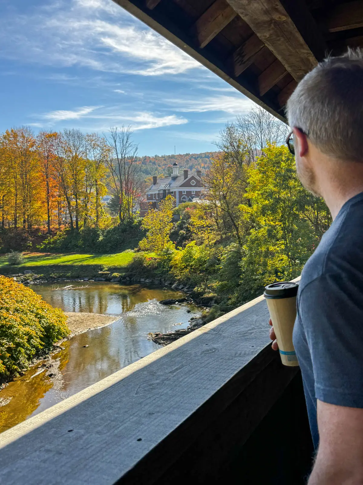 profile of man looking out of a covered bridge in downtown stowe in fall with river below and green and orange trees lining the river.