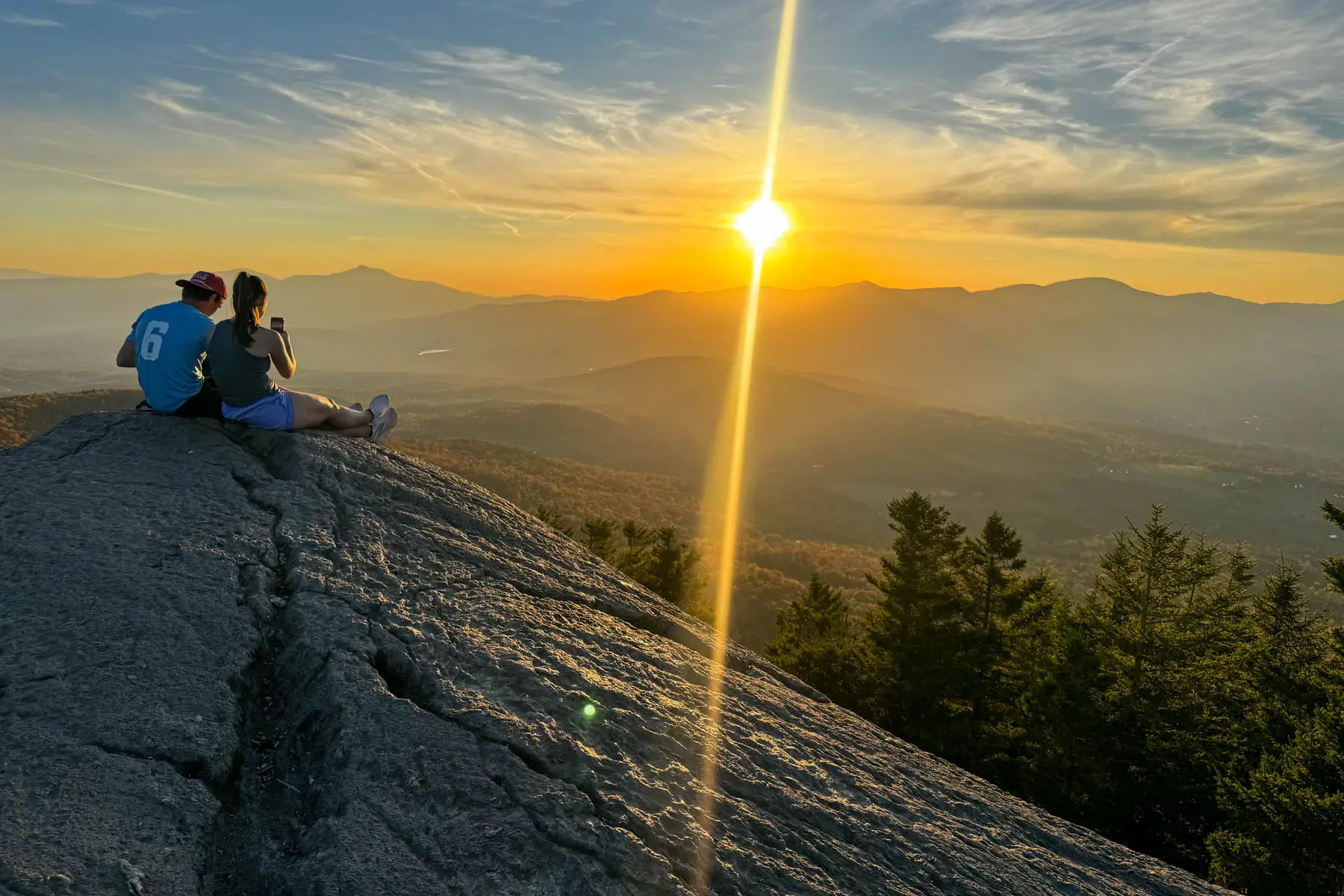 silhouette of two people sitting at top of rock cliff on sunset hike in stowe vermont with golden sun setting over mountain range in distance and orange rays hitting the silhouette of the dark orange colored mountains in fall.