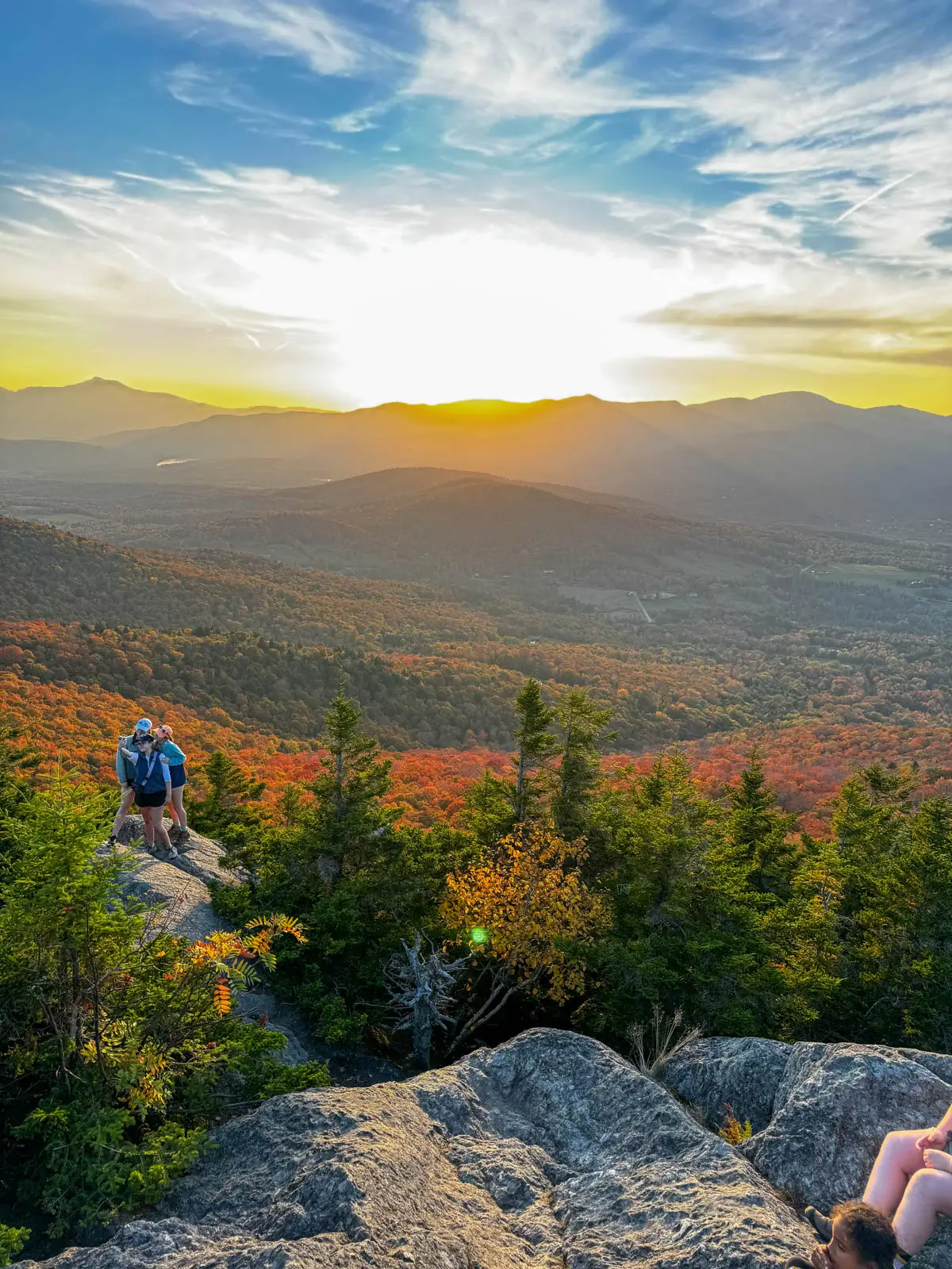 view from rock cliff on sunset hike in stowe vermont with golden sun setting over mountain range in distance and orange rays hitting the silhouette of the dark orange colored mountains in fall.