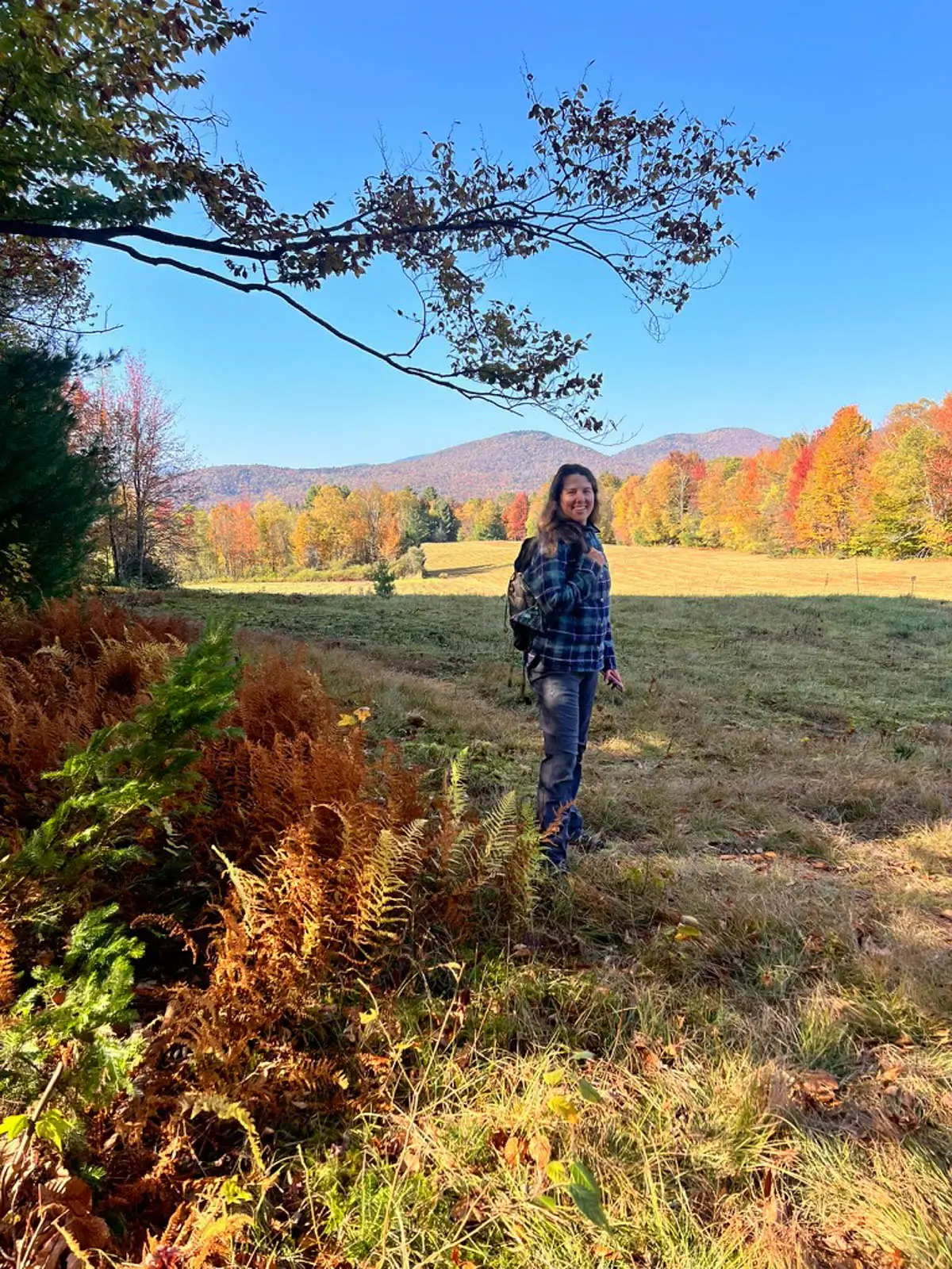 woman standing in view of open meadow on a hike in fall in stowe with hills of orange and green trees in the distance and blue sky above.