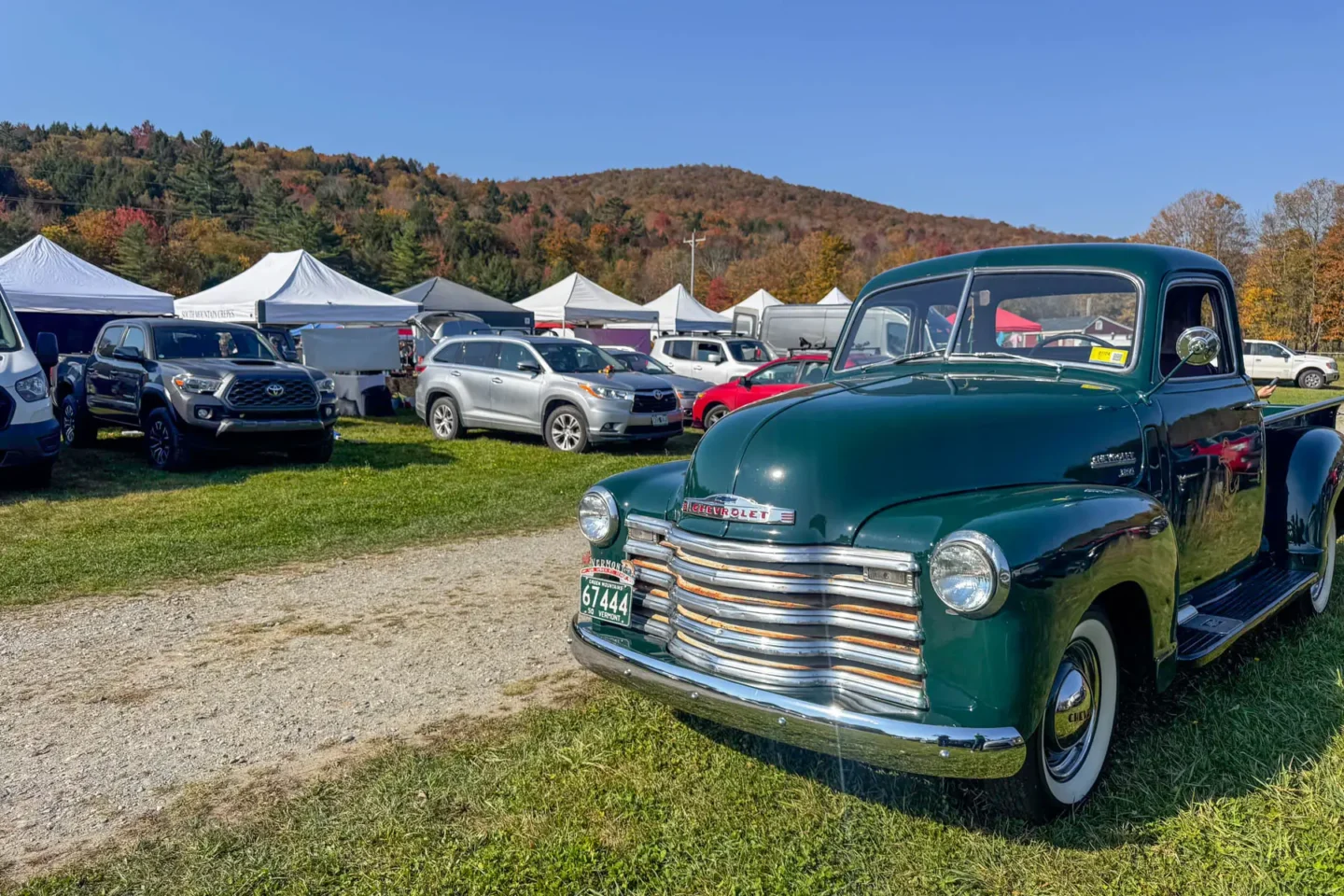 a farmers market in stowe vermont in the fall with white tents set up on a green lawn with an antique forest green car parked in front and an orange hill in the distance.