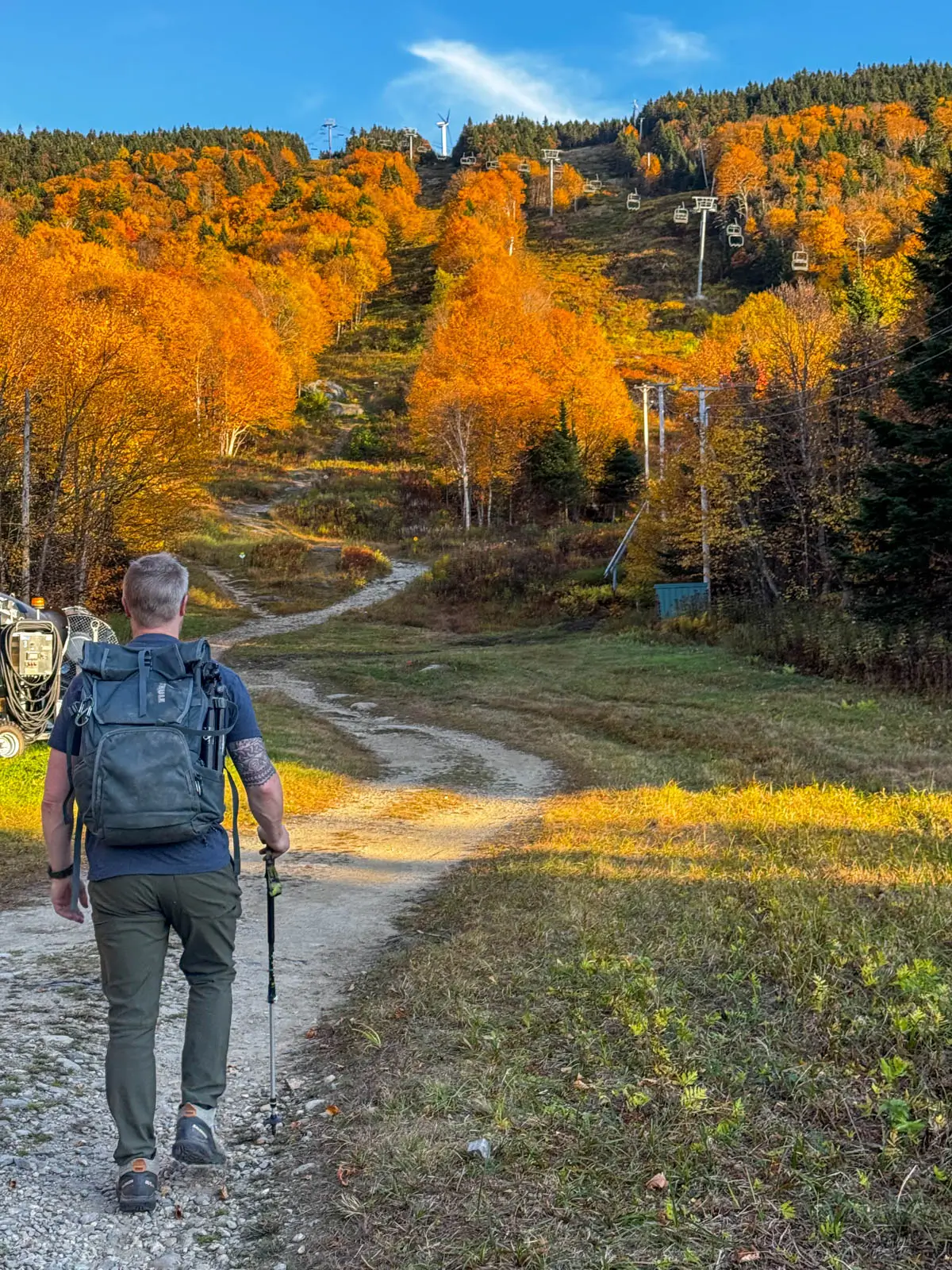 back of man with light blue backpack walking up a winding dirt trail heading into brightly orange colored woods and trees in the fall with a blue sky above.