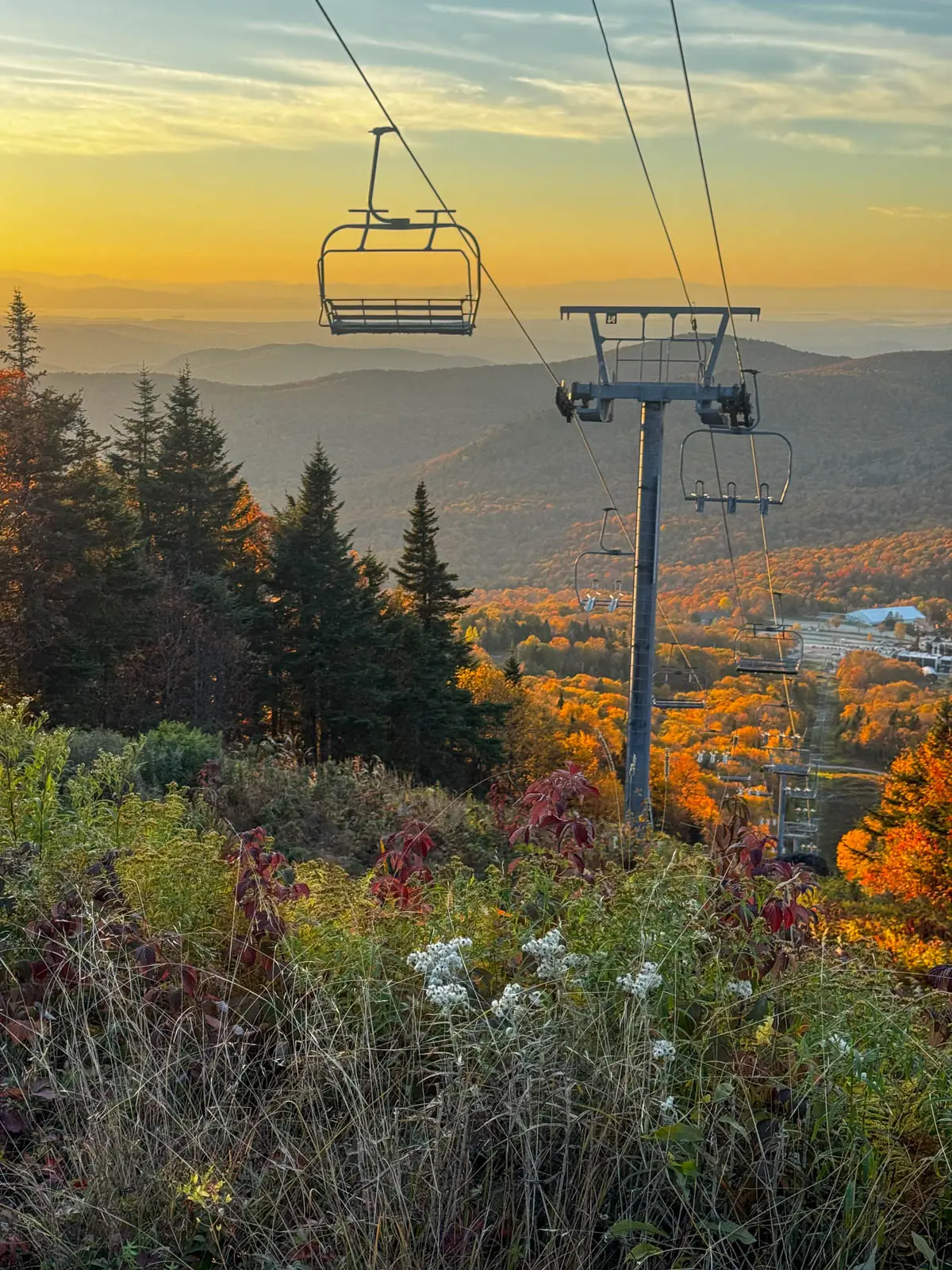 view from a hike at sunset in vermont with a ski lift and silhouette of ski chairs with orange mountains in fall and tall green pine trees in the distance and below.