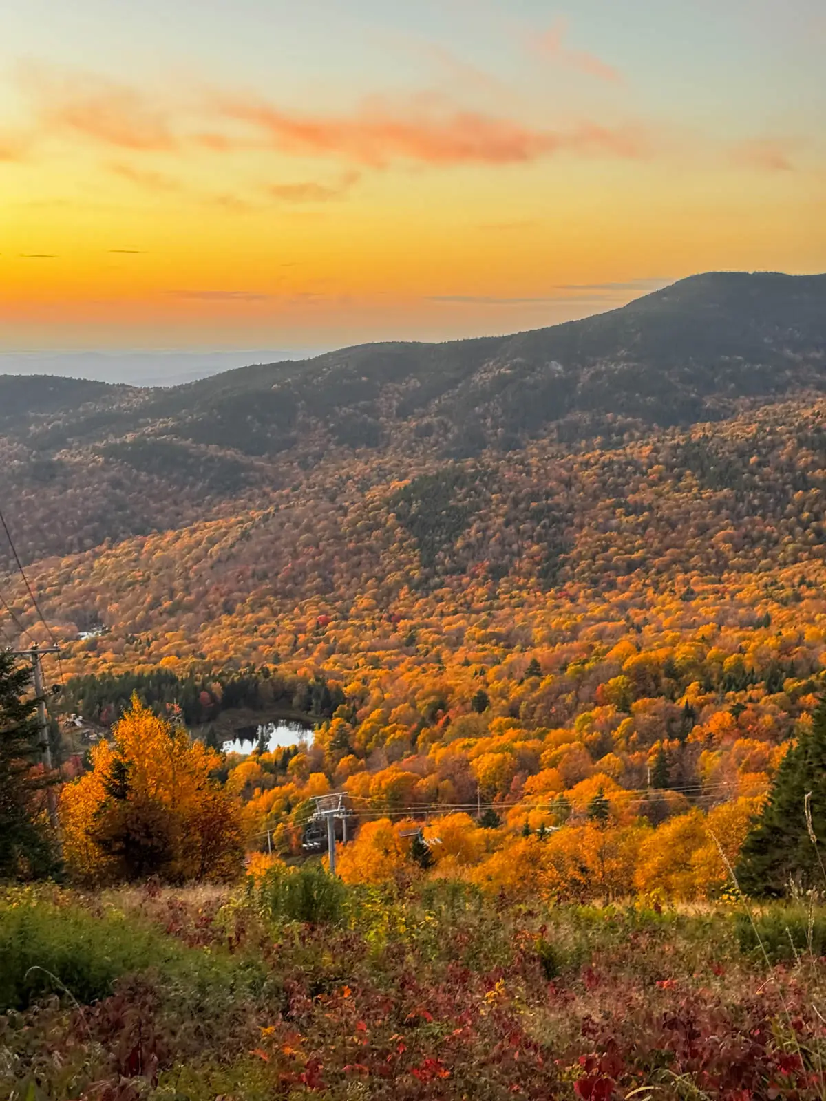 view from top of stowe pinnacle hike in fall at sunset time with view of bright orange colored mountains rolling in the distance and soft orange glow in the sky.