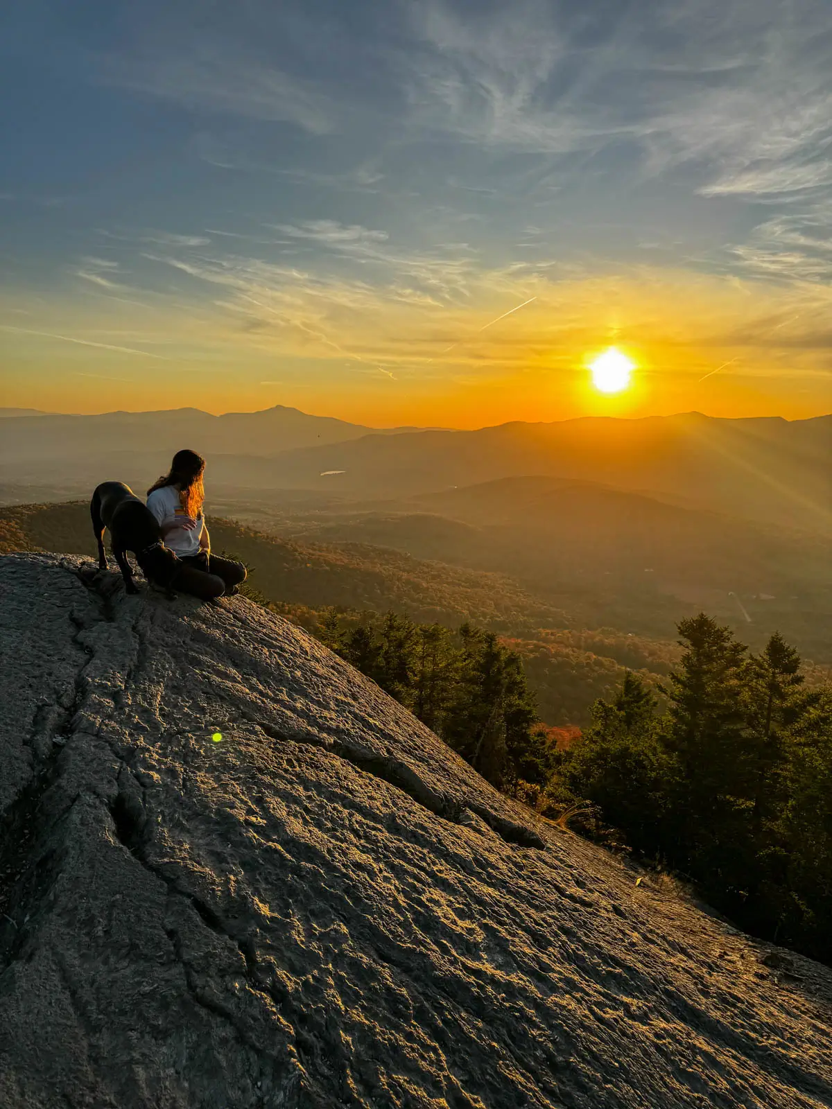 silhouette of woman and dog sitting at top of rock cliff on sunset hike in stowe vermont with golden sun setting over mountain range in distance and orange rays hitting the silhouette of the dark orange colored mountains in fall.