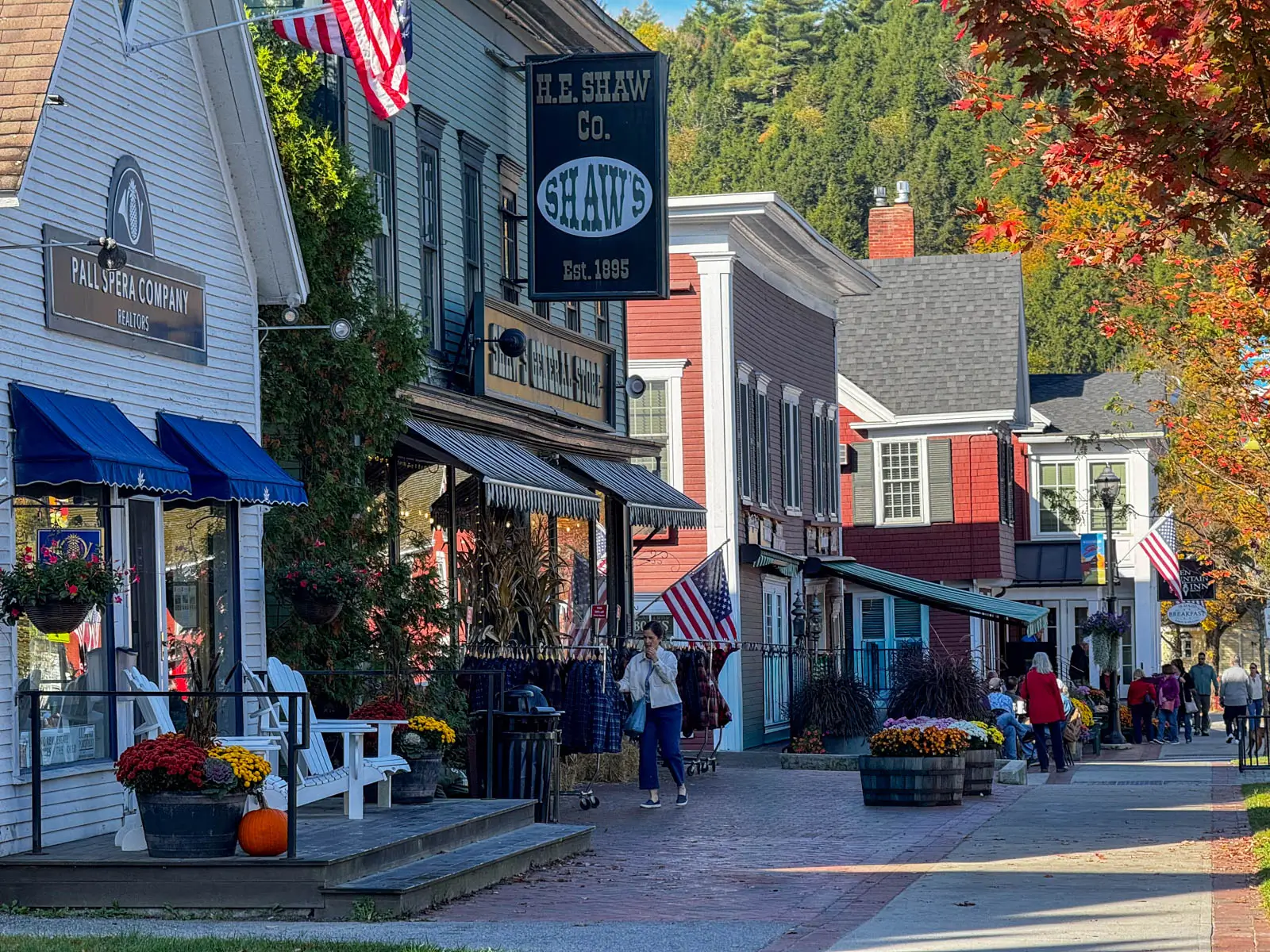 view of downtown stowe at fall with several storefronts lining the street and orange and green hill iin background and orange maple trees liniing the street.