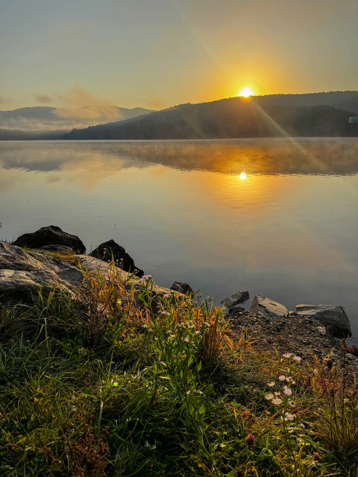 a lake at sunrise in fall with layer of thin fog on the lake and far side of lake lined with dark green pine trees and orange colored trees and a cloudy sky with hint of an orange glow.