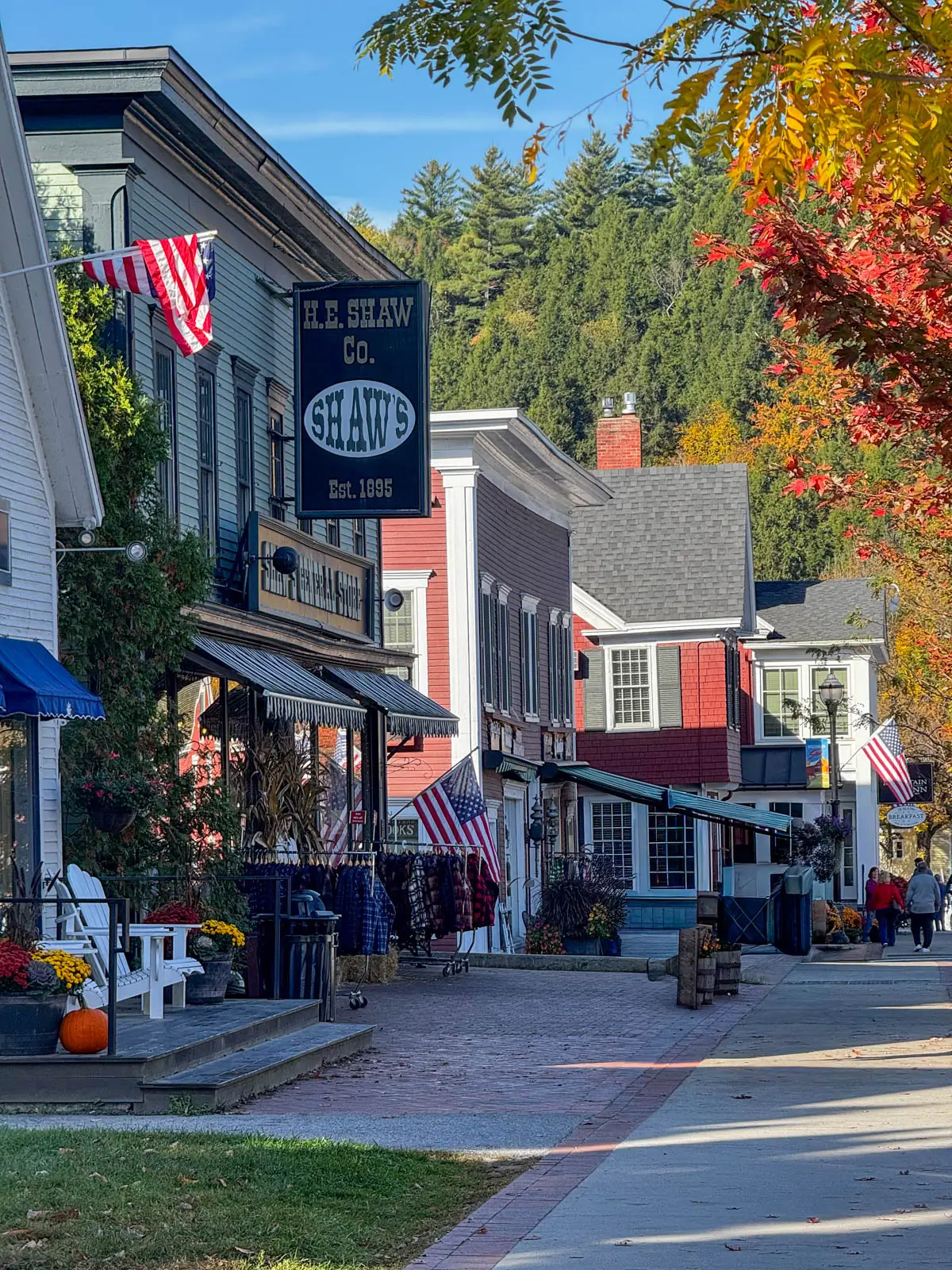 view of downtown stowe at fall with several white and red historic storefronts lining the street and orange and green hill iin background and orange maple trees liniing the street.
