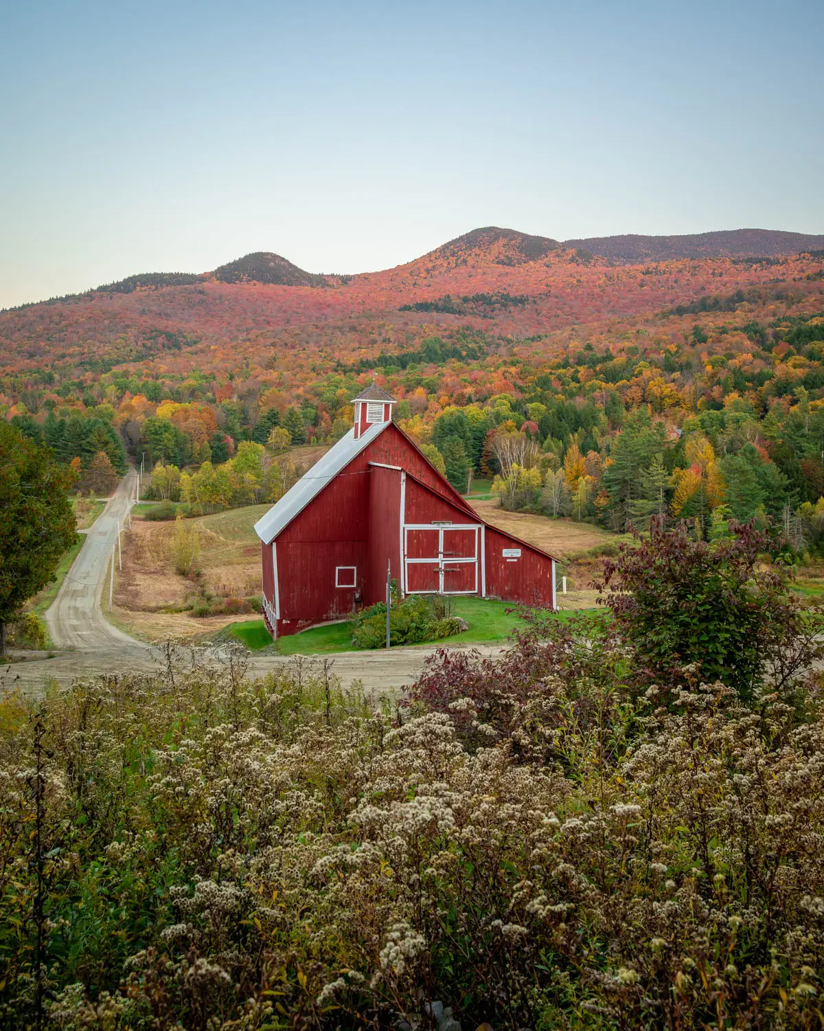 red barn on dirt road in stowe vermont in fall with orange mountain range in distance behind the barn and soft blue sky above.