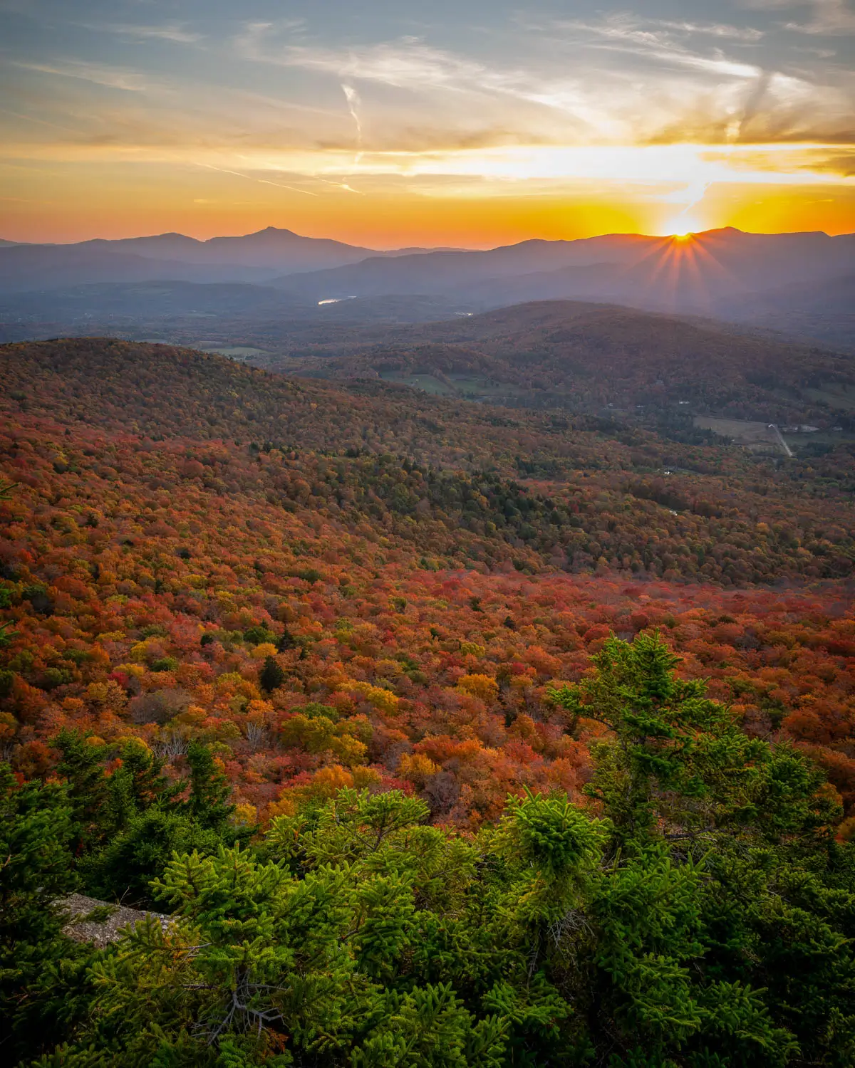 view from top of stowe pinnacle hike in fall at sunset time with view of bright orange colored mountains rolling in the distance and soft orange glow in the sky.