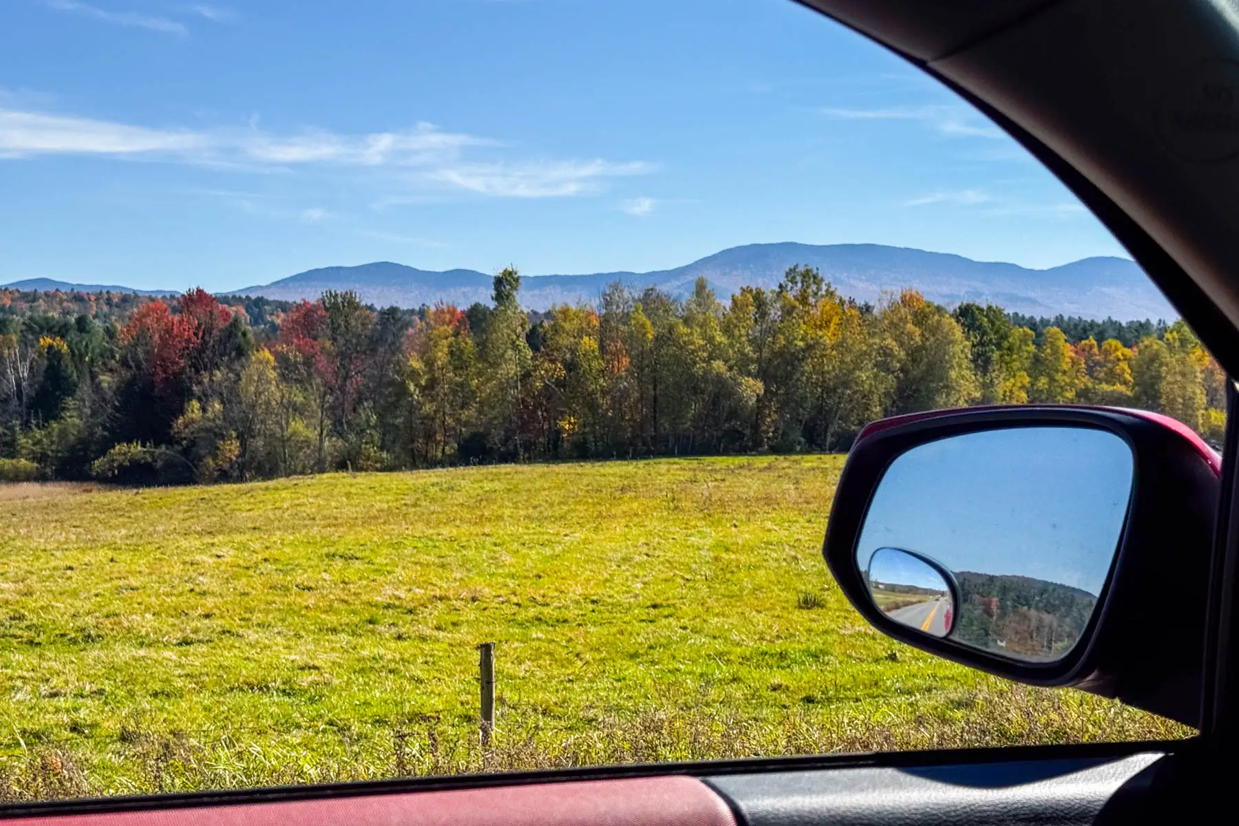 view from driver's side window out of a car at a green meadow lined with fall colored trees and hills in the distance and soft blue sky above.