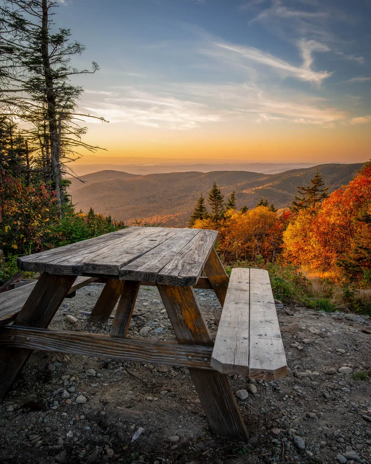 picnic table at top of hill at sunset time in stowe vermont in fall with orange colored trees in distance and golden mountain range and orange sky.