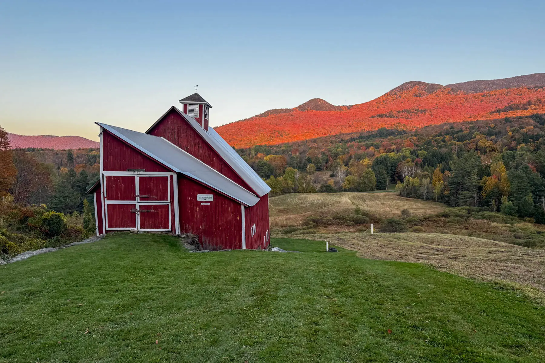 landscape photo of dirt road on left and profile of red barn on the right in the fall with orange and yellow mountain range in the distance.