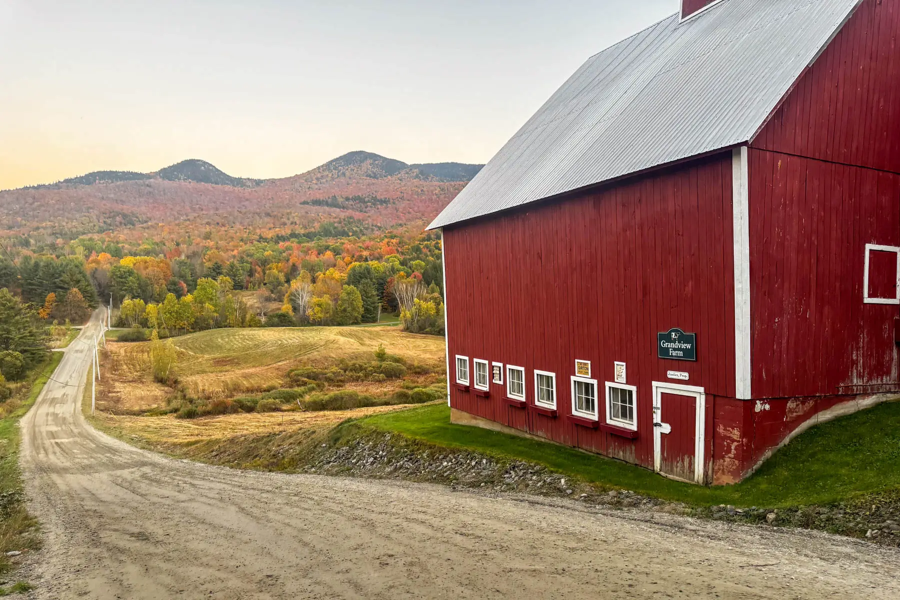 landscape photo of dirt road on left and profile of red barn on the right in the fall with orange and yellow mountain range in the distance.