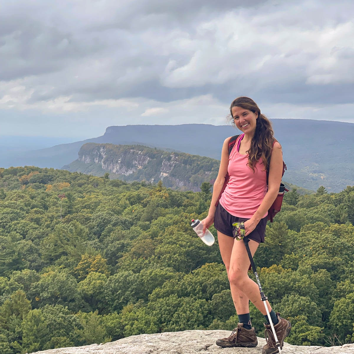 woman in pink tank top and maroon shorts on top of ridge in new paltz on a hike in mohonk preserve with green ridge and trees in background.
