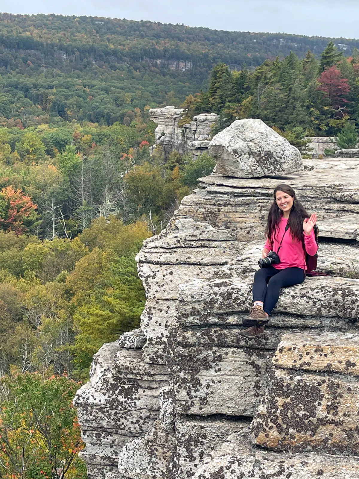 woman in pink shirt smiling and sitting on rocky cliff face with rolling hills and autumn colored trees behind her.