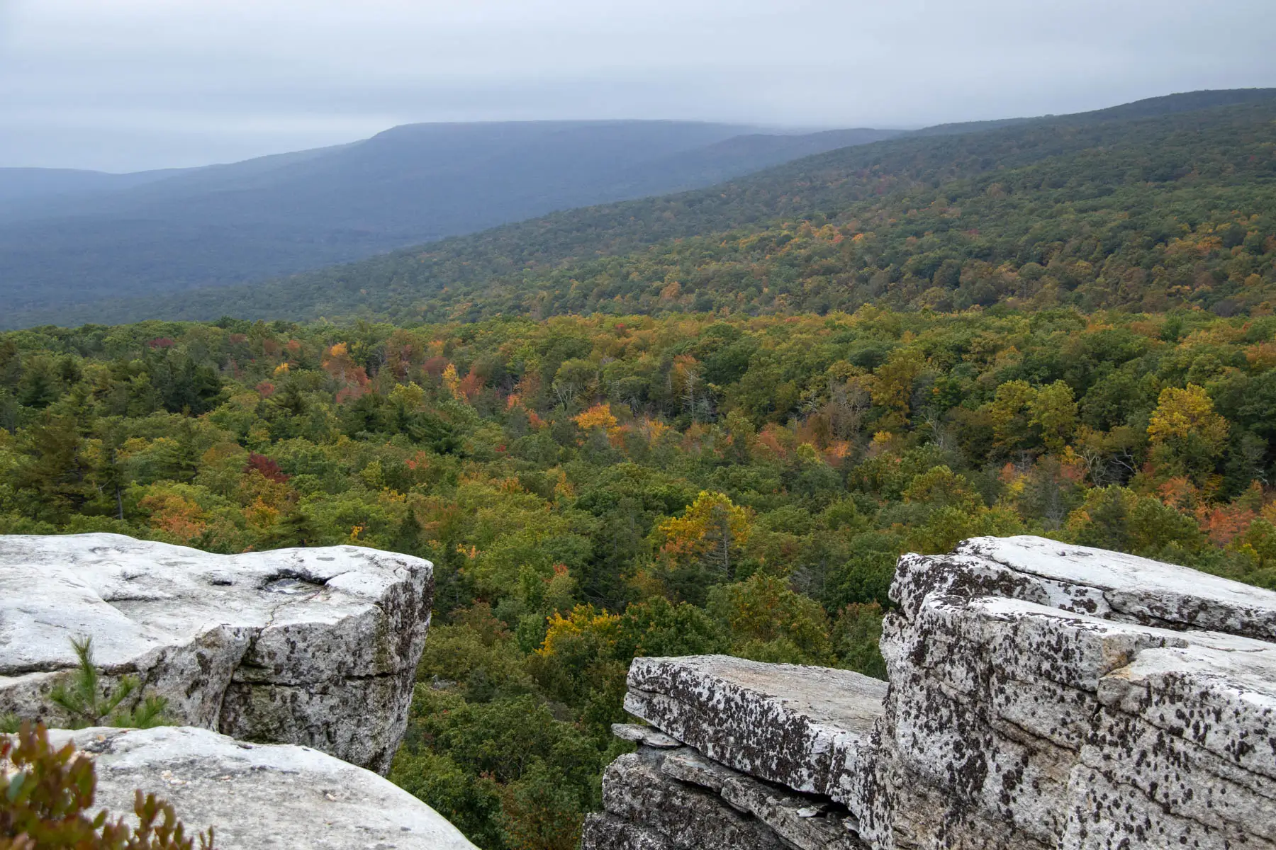 view from a rocky ridge looking out over rolling dark green mountains in late summer with a darker sky and layer of clouds.