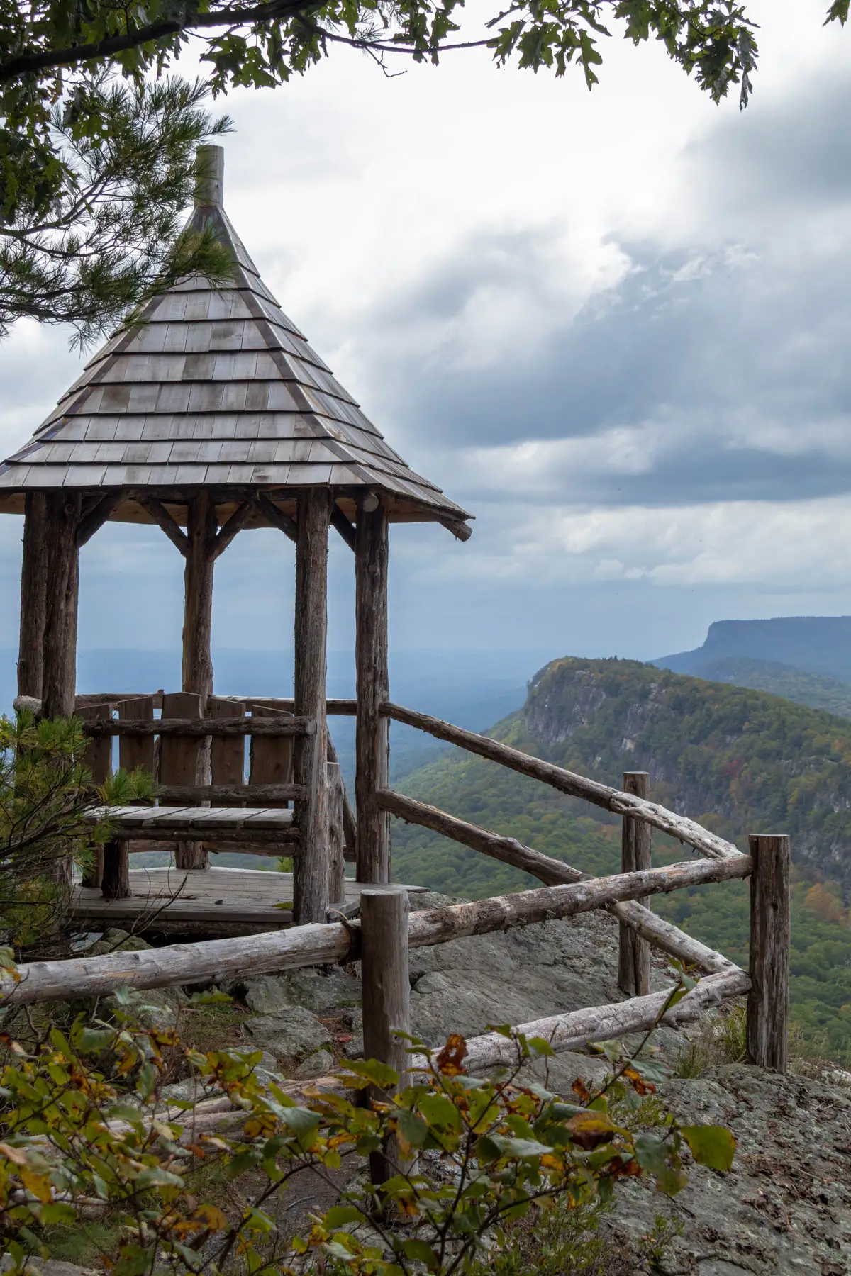 small wooden tower with a bench inside and wooden railing leading up to it at top of hike in mohonk preserve with a mountain ridge behind it in the distance.