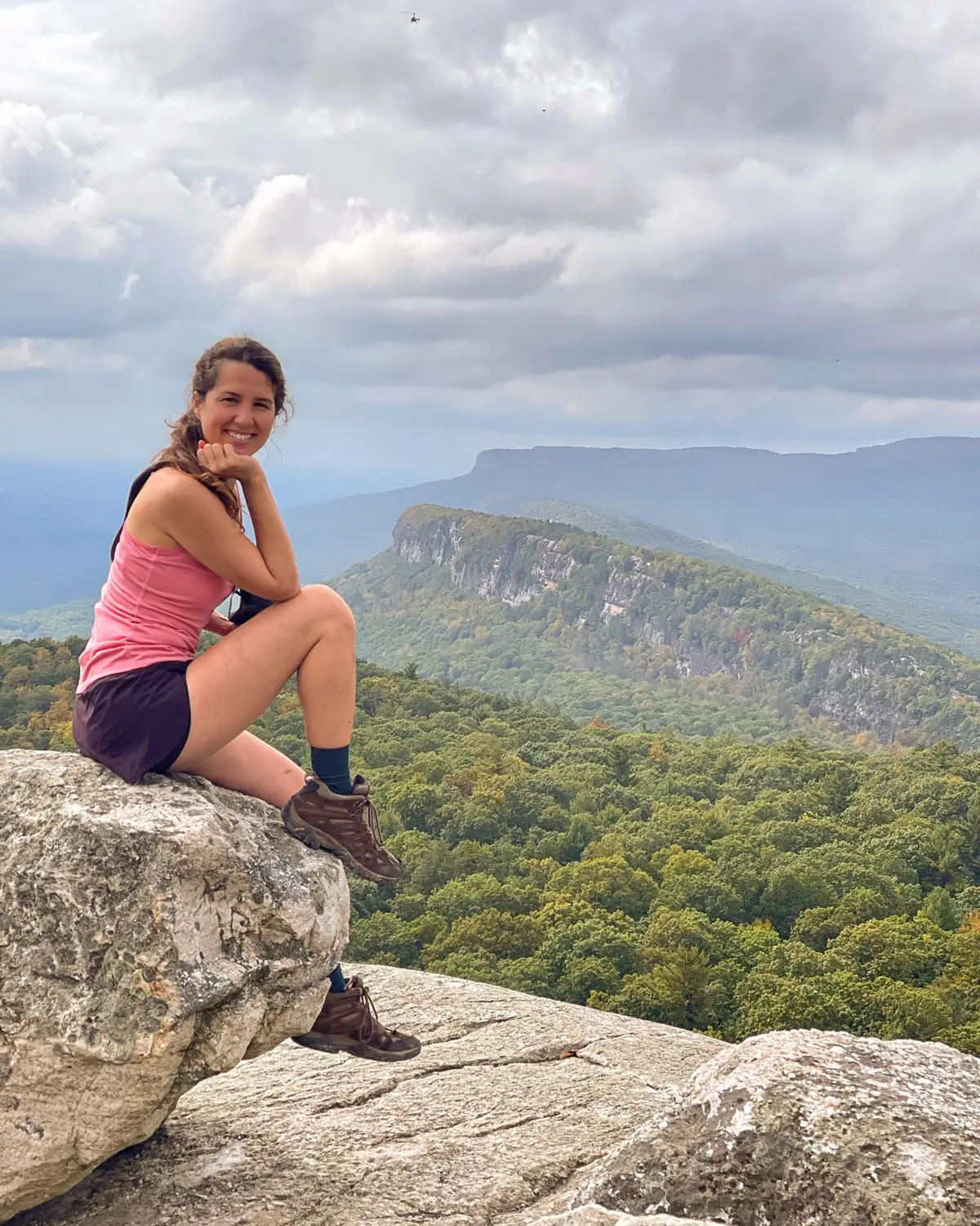 woman in pink tank top and maroon shorts on top of ridge in new paltz on a hike in mohonk preserve with green ridge and trees in background.
