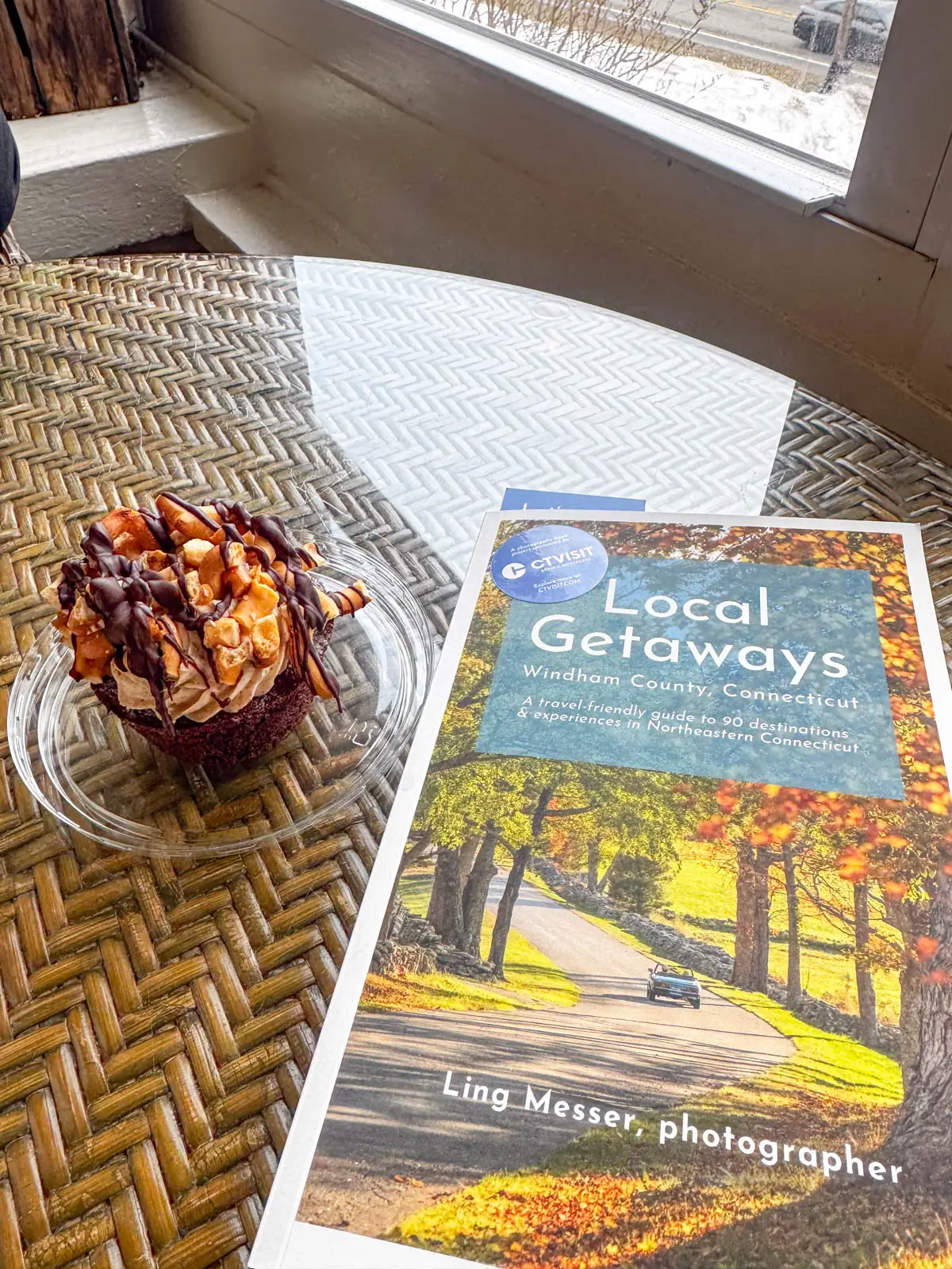 a chocolate cupcake on glass tabletop with a small book net to it on right titled "local getaways" depicting a country road in connecticut.
