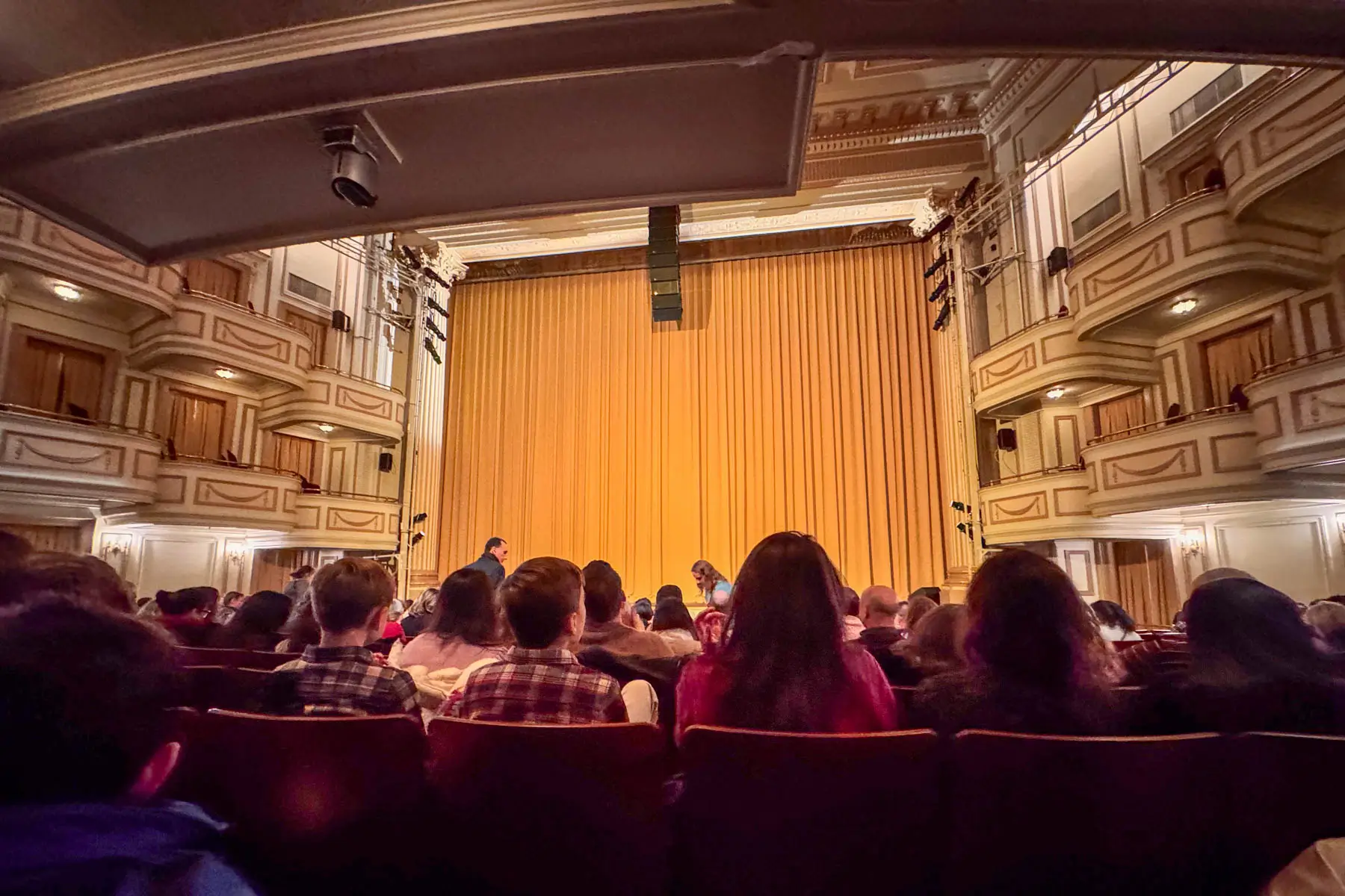 inside shubert theater with rows of reds seats with back of peoples heads facing the stage with long gold curtain.