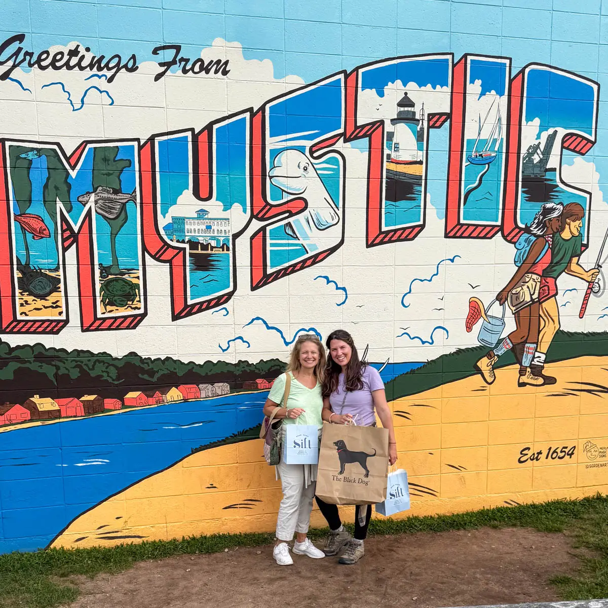a brunette and blonde woman standing in front of a wall mural outside with the word "mystic' painted on it with a beach theme.