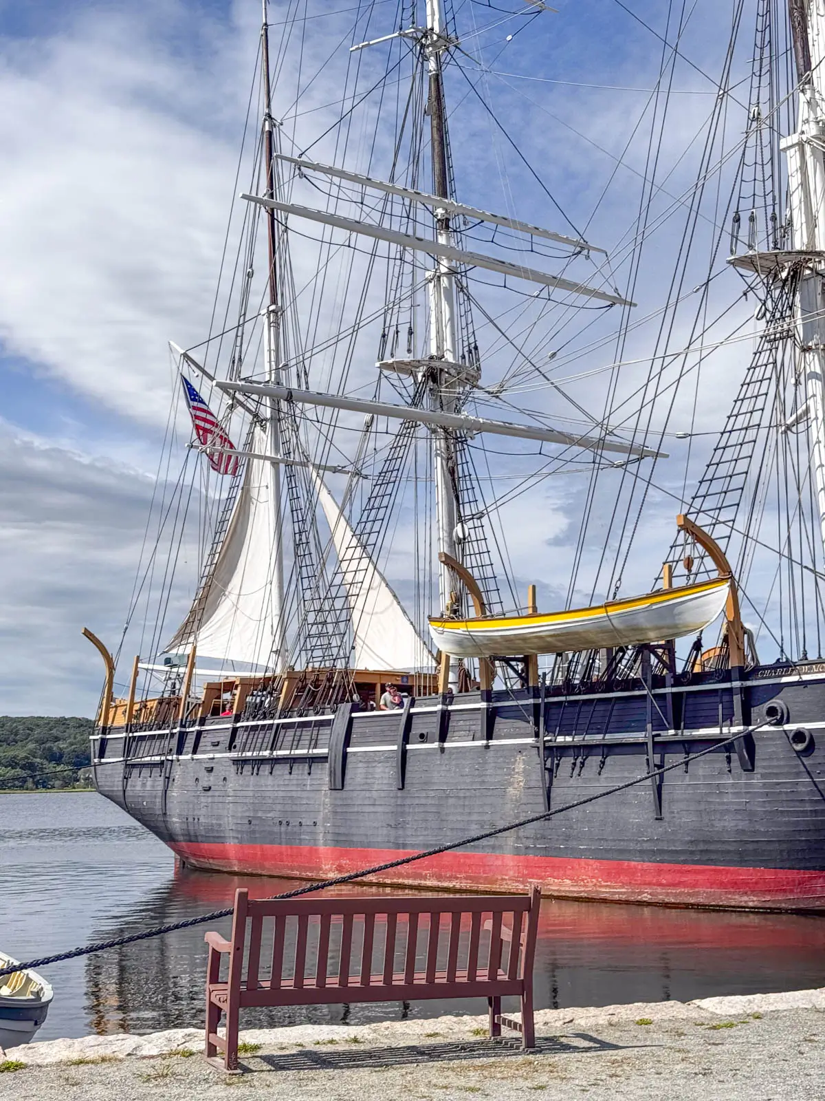 an old ship with huge white masts and a black wooden base with red trim on bottom in a harbor in Mystic.
