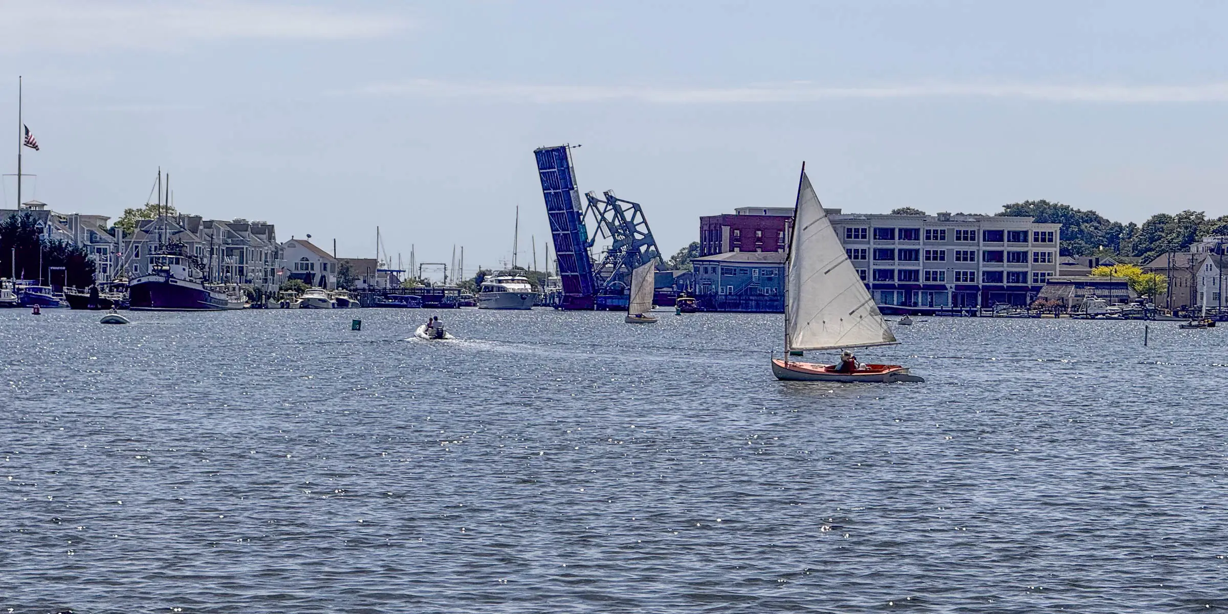 white sail boat in blue water with a drawbridge in the distance.