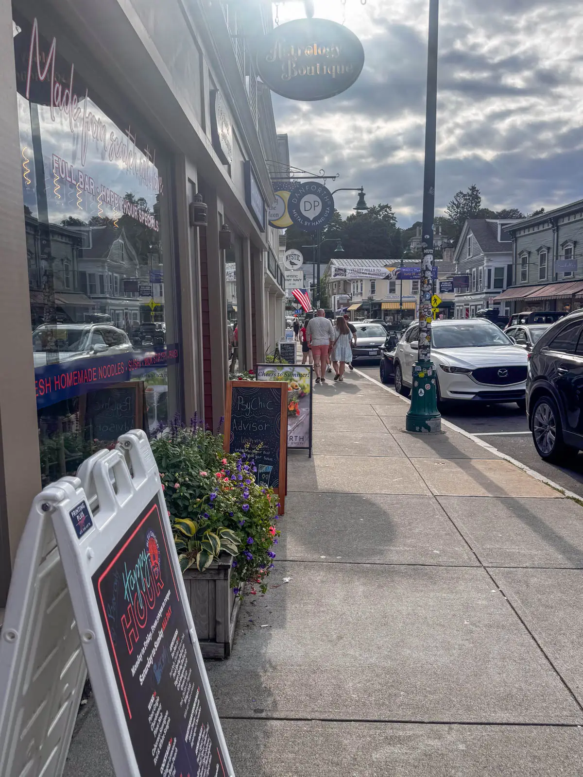 a street in downtown Mystic with shops lining the left side and people walking down the sidewalk with a hazy sky on a summer day.