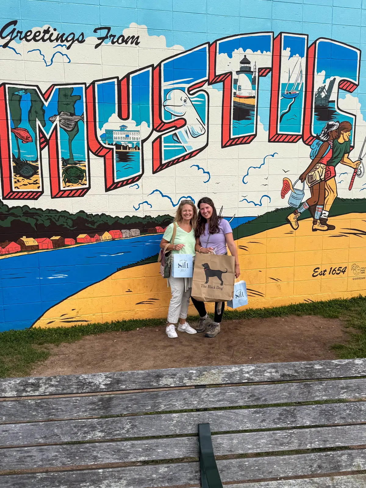 a brunette woman and blonde woman standing next to each other smiling in front of a wall mural that says the word "mystic" with paintings of a beach below it.