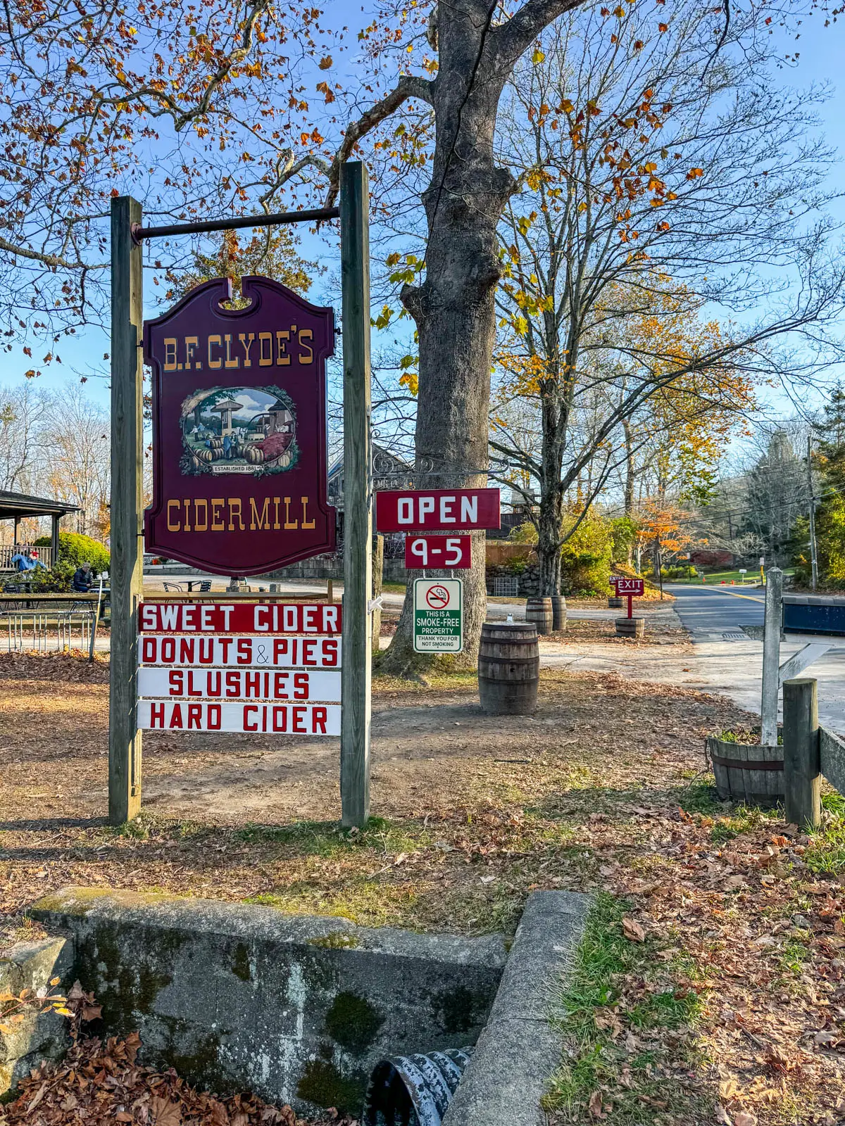 red sign in front of a cider mill with "B.F. Clyde's Cider Mill" written on it in gold and a soft blue sky above and orange colored leaves and trees in background.