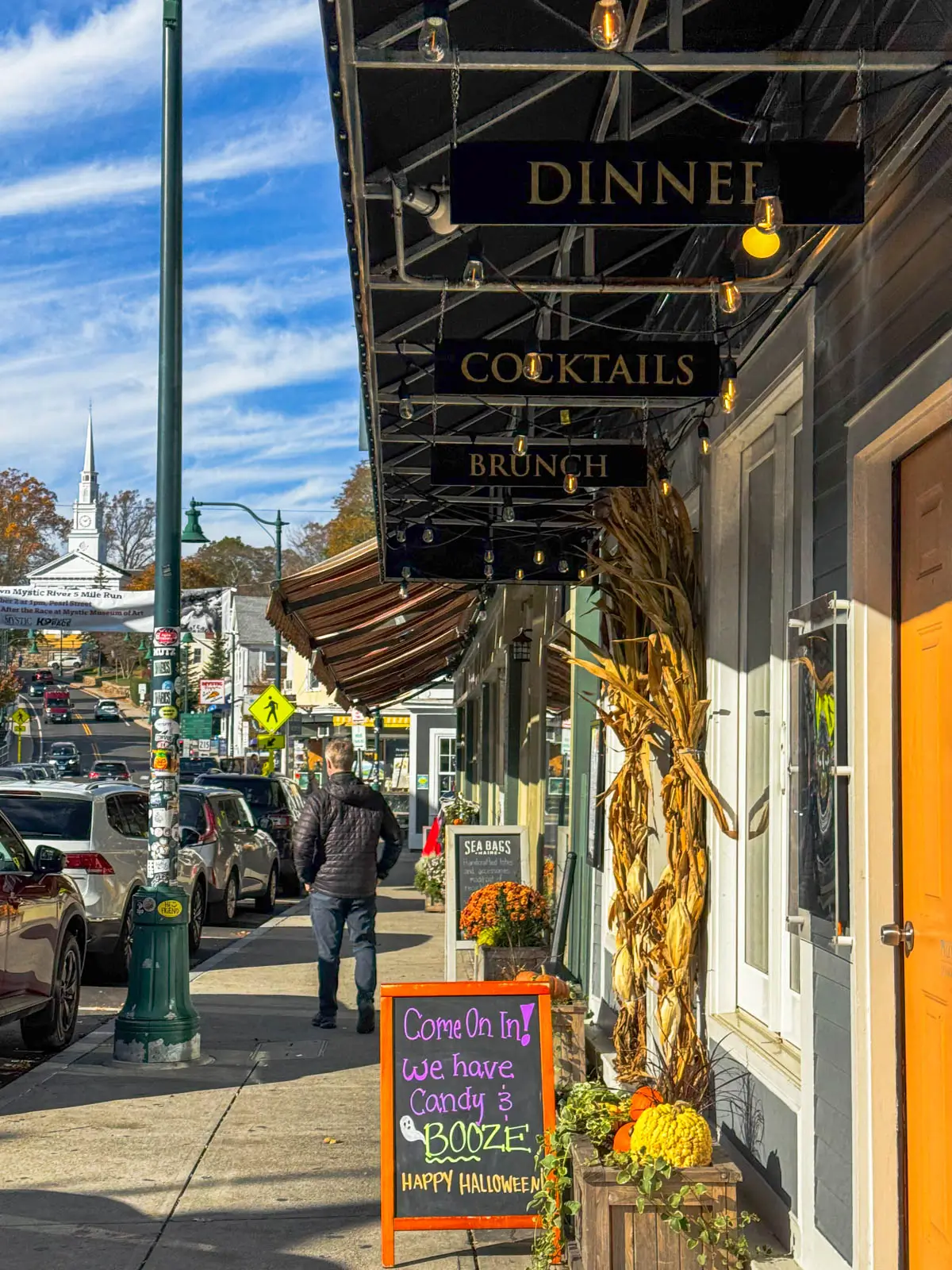 back of blonde man in black fall jacket and long pants walking along a downtown street in fall with mums, corn stalks, and other fall decorations and an orange sign lining storefronts.