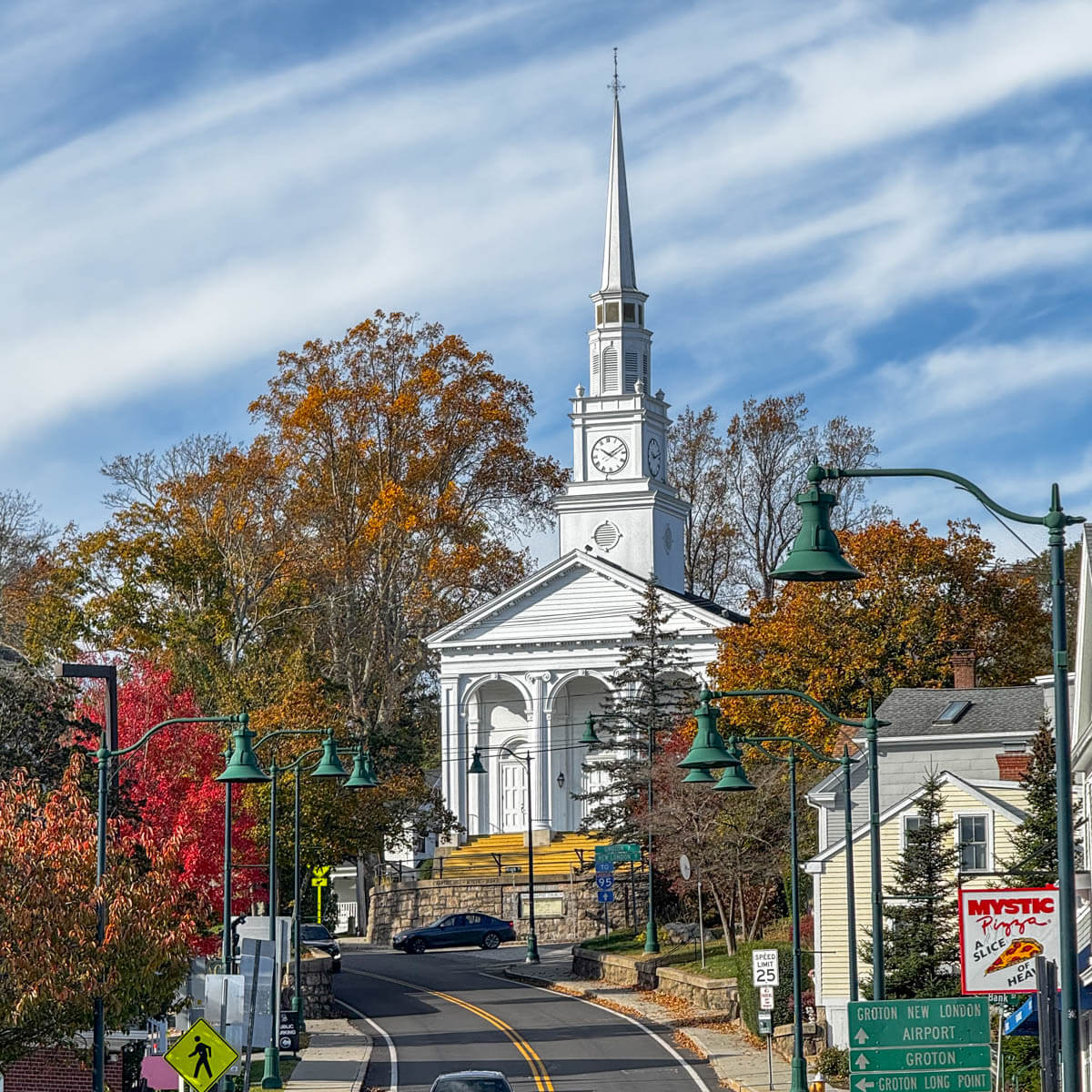 white steeple church atop a hill with a soft blue sky background, white whispy clouds, and orange and red colored trees surrounding the base of the church.