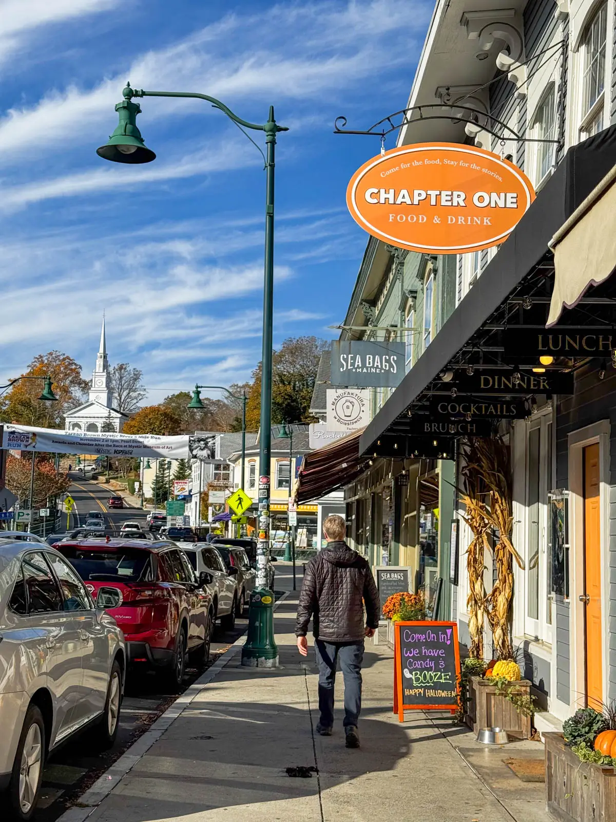 back of blonde man in black fall jacket and long pants walking along a downtown street in fall with mums lining storefronts and an orange sign hanging in top right corder and deep blue sky with wispy clouds above.