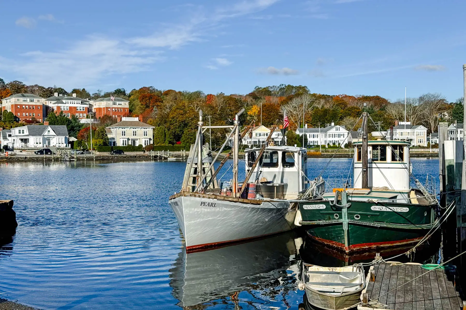 two tug boats (one white and one forest green) in dark blue water with autumn colored trees lining the background on the land and soft blue sky above.