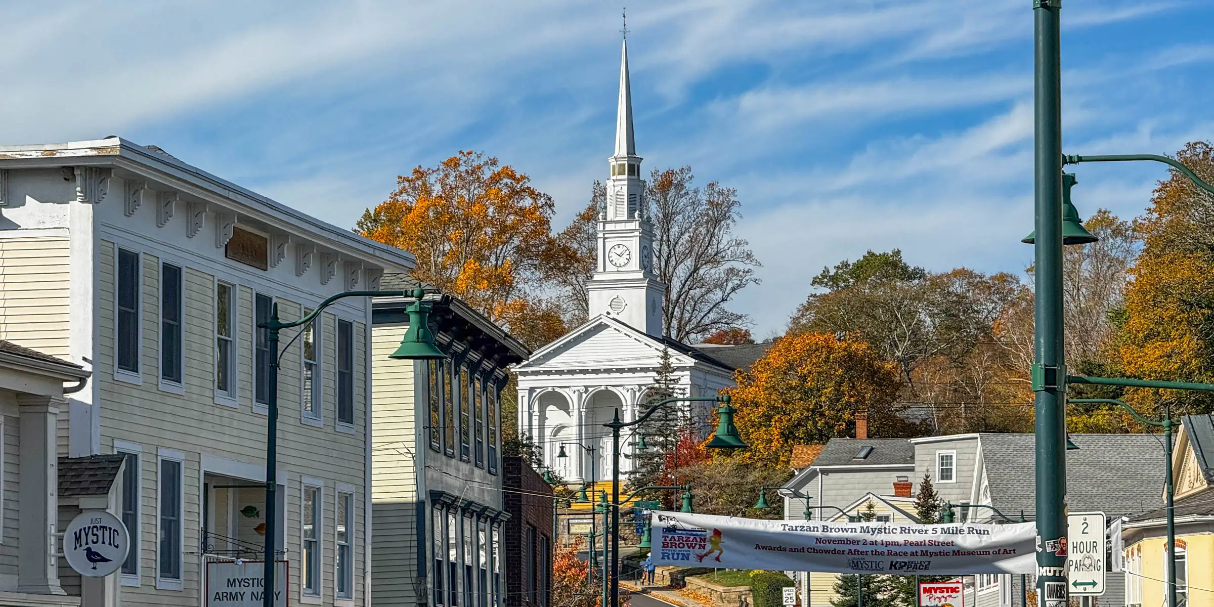wide landscape photo of white steeple church high on hill on a downtown street surrounded by orange. red, and brown colored trees in fall with a soft blue sky above and white wispy clouds.