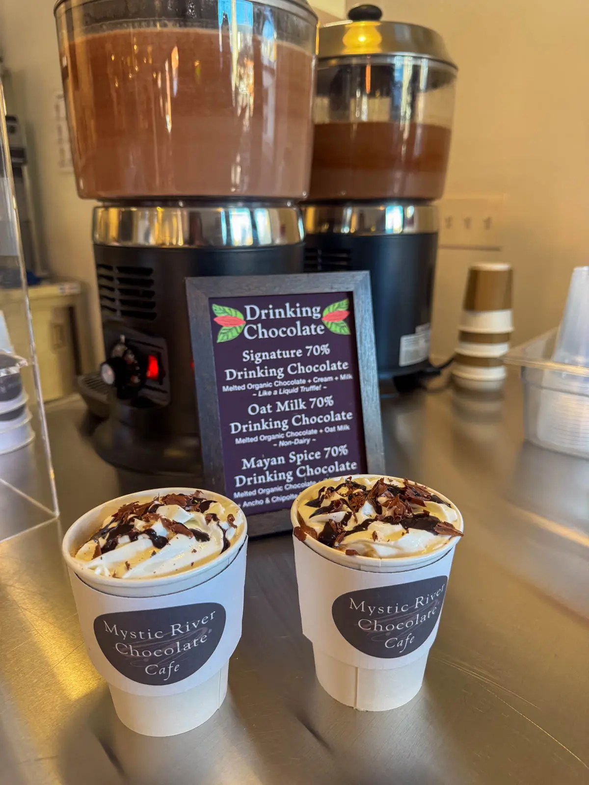 two white paper cups on a coounter with "Mystic River Chocolate" writing in brown filled with hot chocolate and topped with whipped cream and chocolate sauce.